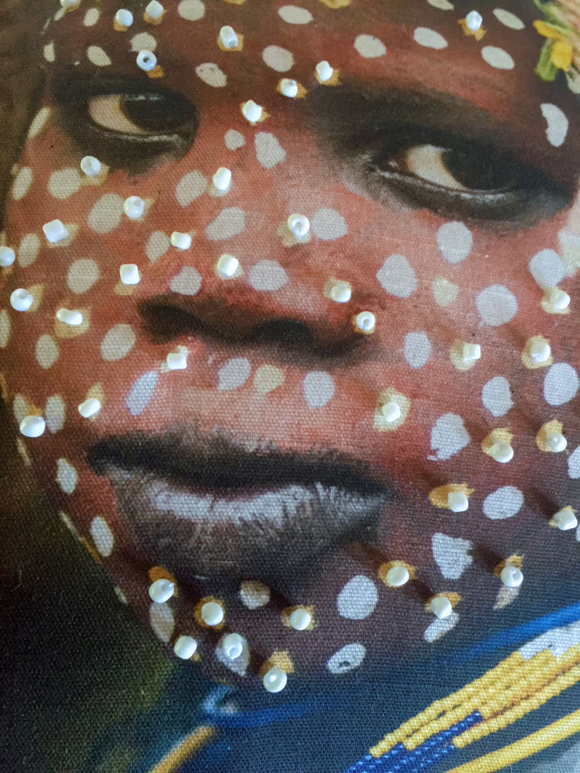 Close-up of a person’s face with brown skin, adorned with white painted dots and small, round beads attached to the surface. The person gazes to the side with a calm expression. Blue and yellow fabric is visible at the bottom edge.