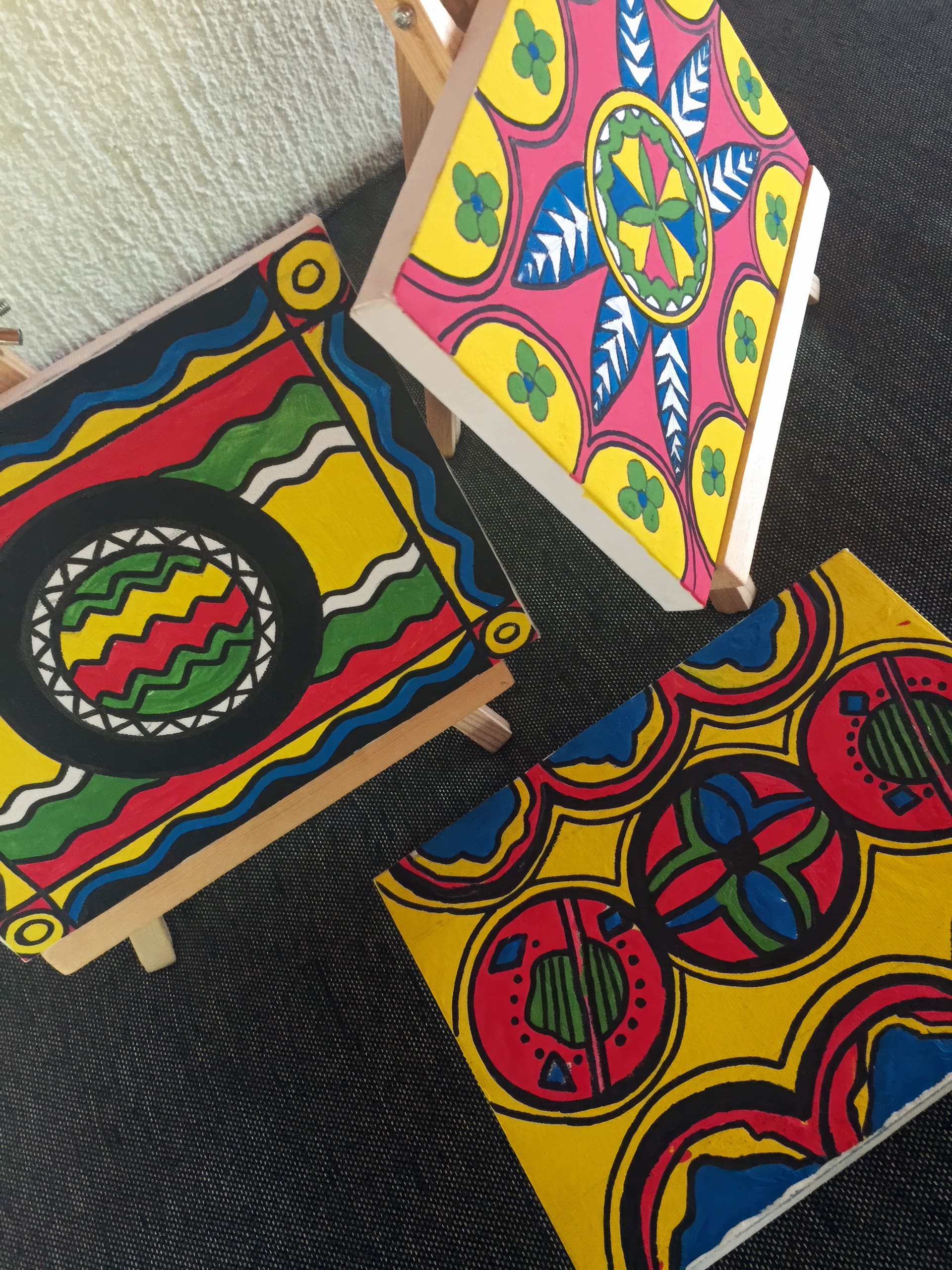 Three brightly coloured paintings with bold, geometric patterns and vibrant red, yellow, green, blue, and black shapes are displayed on small wooden easels, resting on a dark-textured surface near a light-coloured wall.