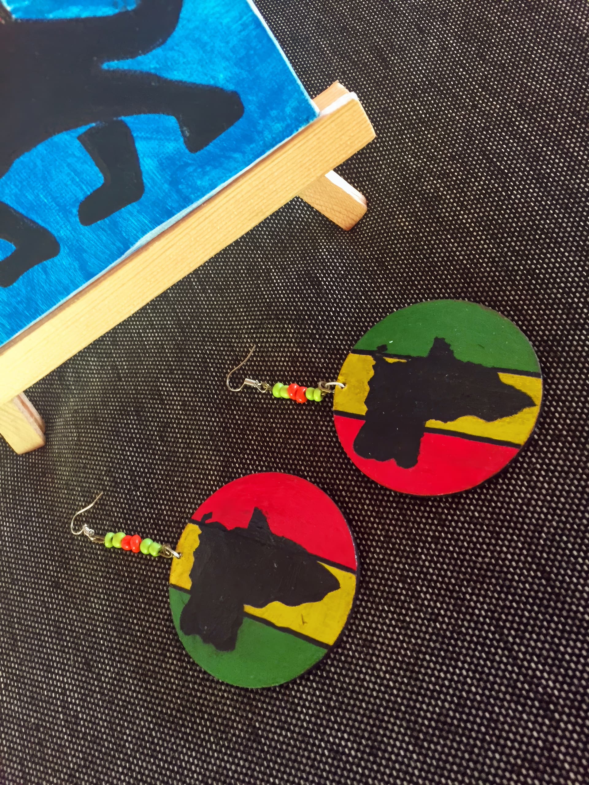 Two round earrings lie on a dark textured surface. Each earring has red, yellow, and green stripes with a black Africa map silhouette. Above the circles are small red, yellow, green, and black beads. Part of a colourful painting on a mini easel is visible above them.