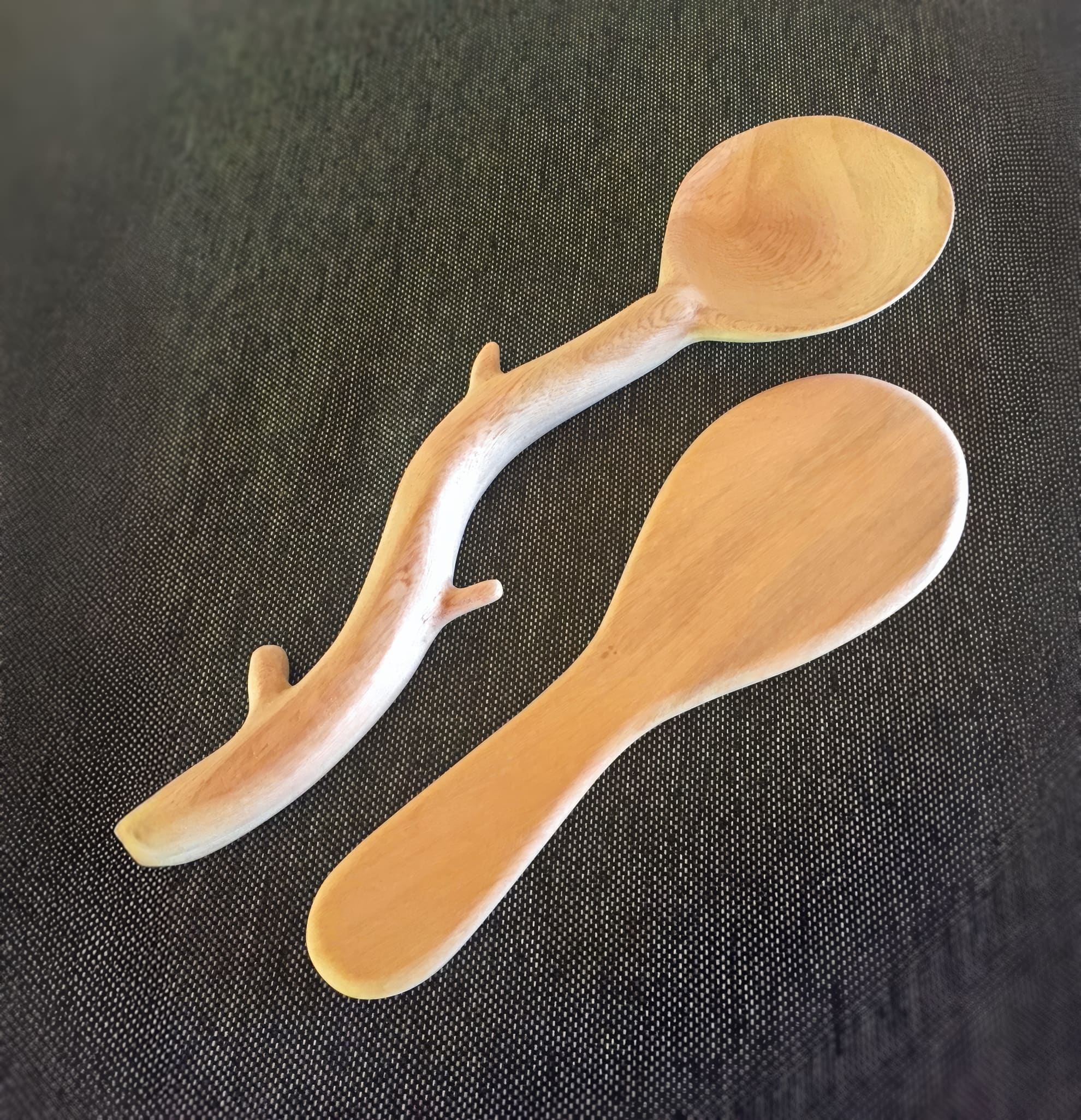 Two wooden spoons lie on a dark textured surface. One spoon has a smooth, conventional shape; the other features a unique branch-like handle with natural protrusions, contrasting traditional and artistic designs.
