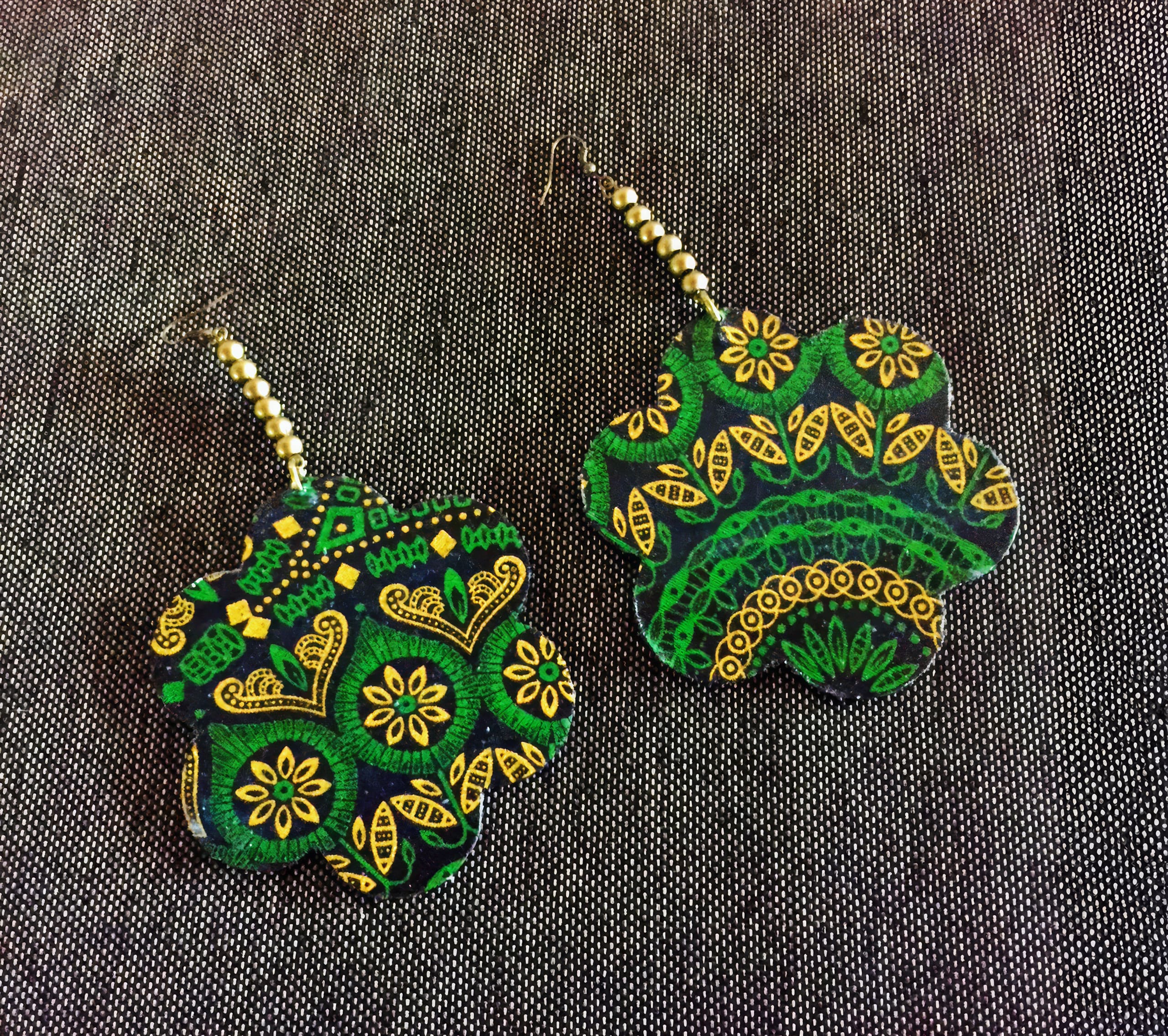 A pair of large, flower-shaped earrings with intricate yellow and green patterns, including flowers, leaves, and geometric lines, lie on a textured dark grey fabric background. The earrings have beaded hooks with alternating gold and black beads.