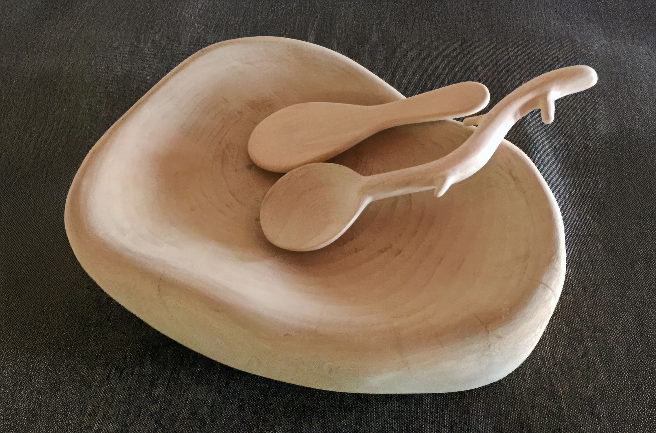 A smooth, light-coloured wooden bowl with an organic, asymmetrical shape sits on a dark textured surface. Two matching wooden utensils rest inside, one of which has a unique handle resembling an animal or branch with protruding elements.