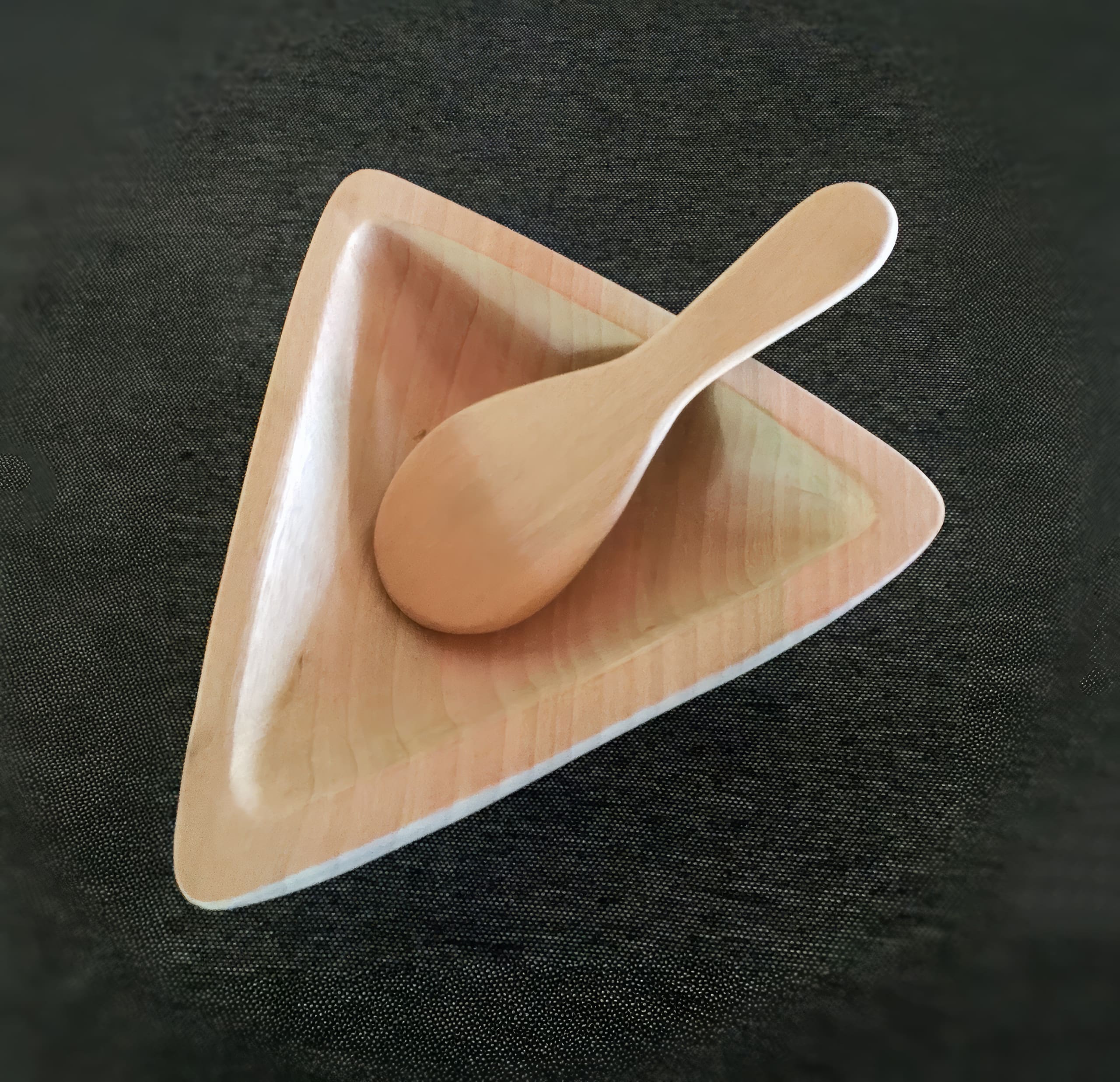 A light wooden spoon rests inside a triangular, shallow wooden bowl. Both items are placed on a dark, textured surface, creating a contrast between the light-coloured wood and the dark background.