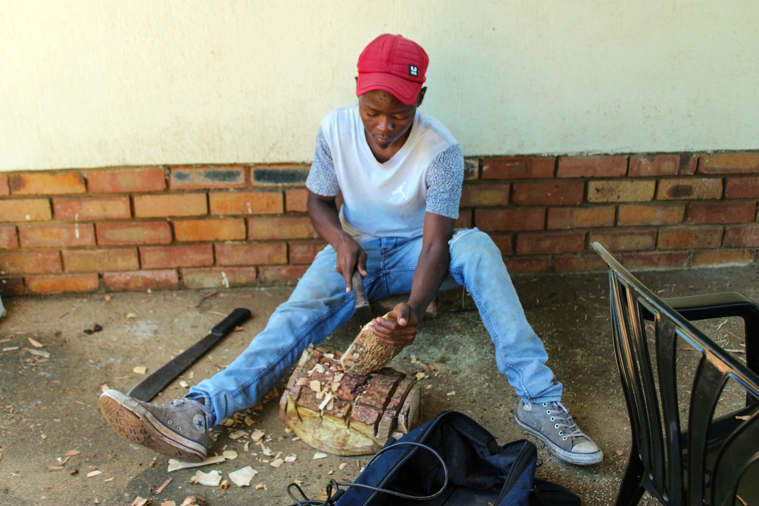 A person in a red cap, white shirt, and blue jeans sits on the ground, carving wood with a knife. Wood shavings and tools are scattered around. They are in front of a brick wall near a black plastic chair and a rucksack.