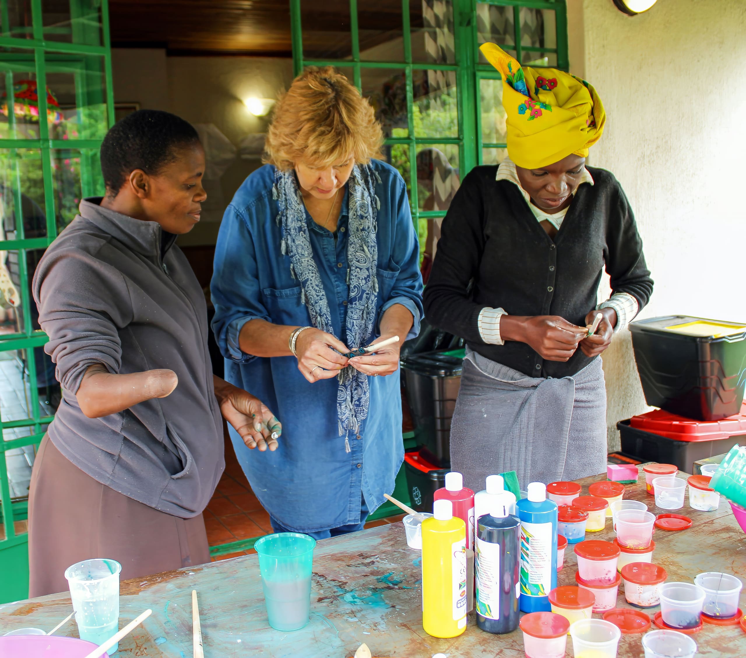 Three women stand around a table covered with colourful paints and art supplies. One woman has a limb difference, another wears a blue scarf, and the third wears a yellow headwrap. They focus on small objects in their hands, engaging in an arts and crafts activity.