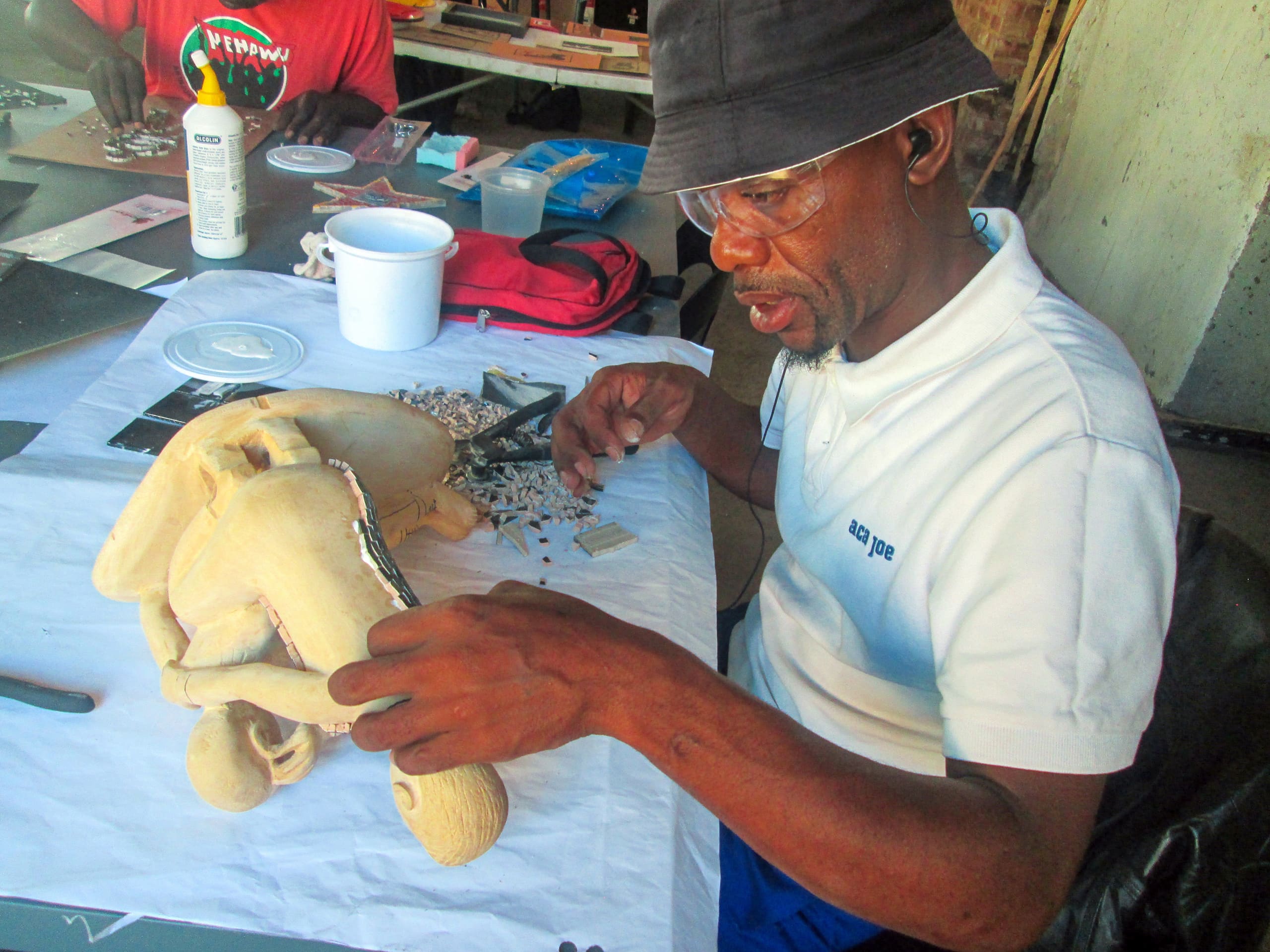 A man in a white shirt, glasses, and a black bucket hat uses tweezers to carefully place small mosaic tiles on a yellow sculpture shaped like a motorbike. He sits at a table covered with art supplies and tools, concentrating on his detailed work.