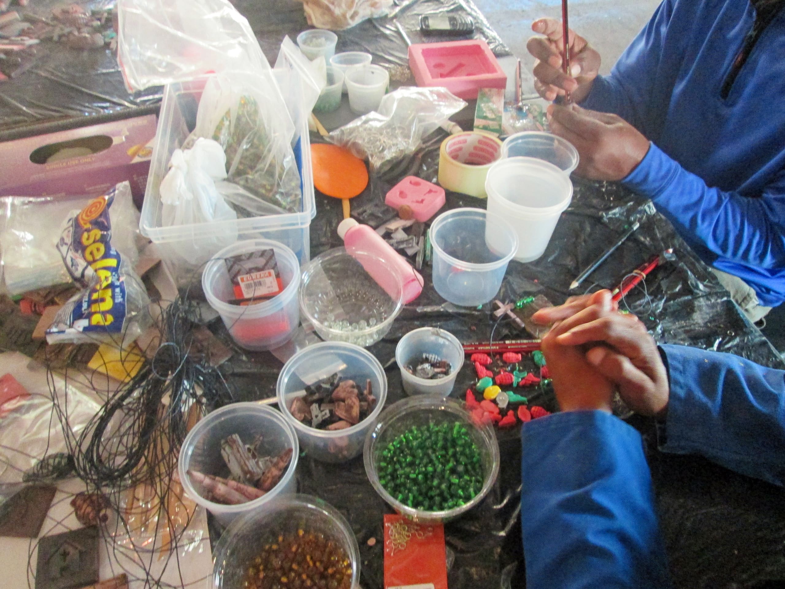 Two people sit at a cluttered table making crafts. Containers with beads, stones, and craft tools are spread out. Hands are visible shaping clay or beads. The table is covered with a black plastic sheet and various colourful supplies and moulds are visible.