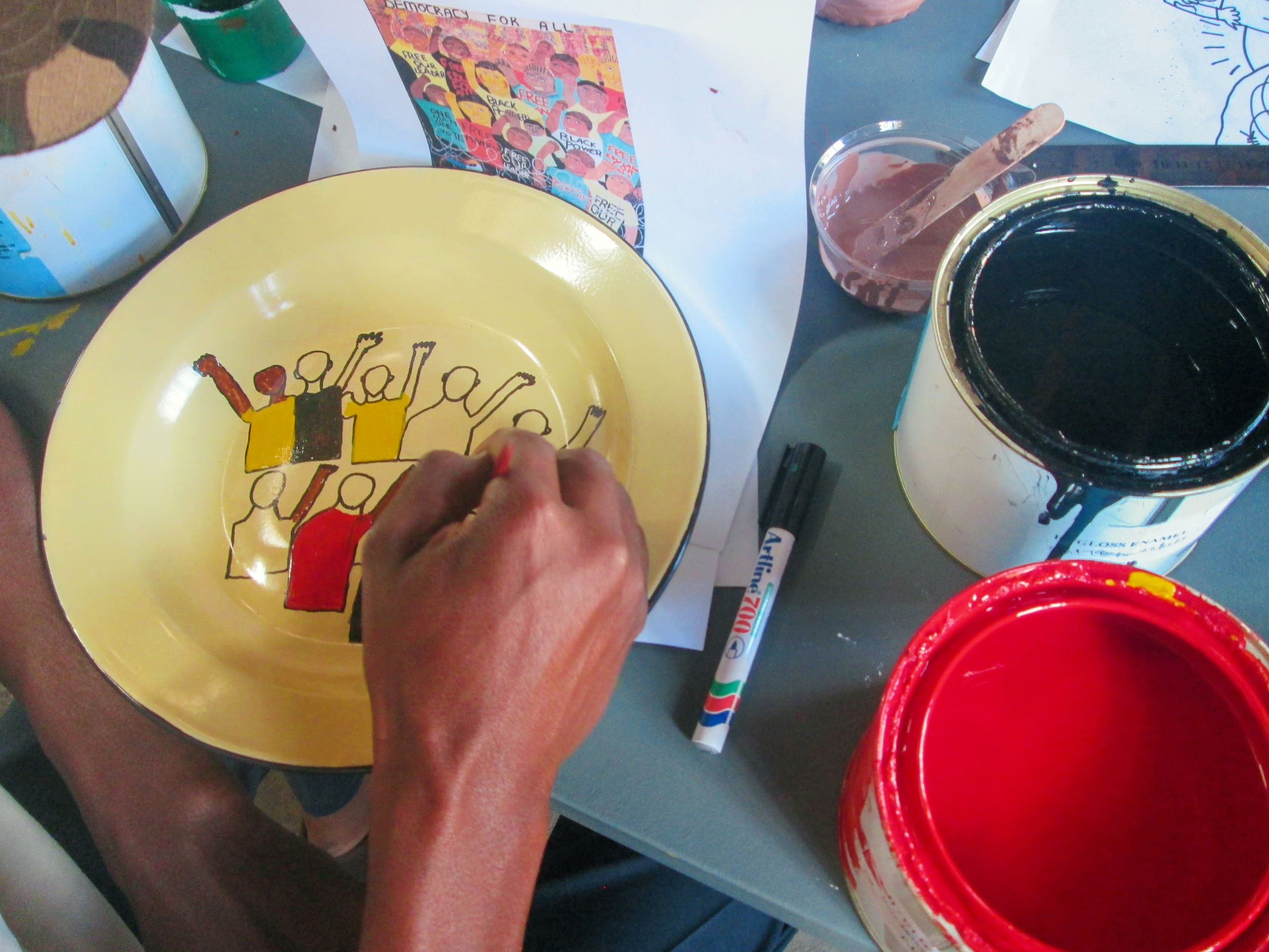 A person paints a design of stylised, raised fists on a yellow plate using black, red, and brown paints. Paint tins, a marker, a paintbrush, and a reference drawing are scattered on the grey table. The scene suggests an art or activism project.