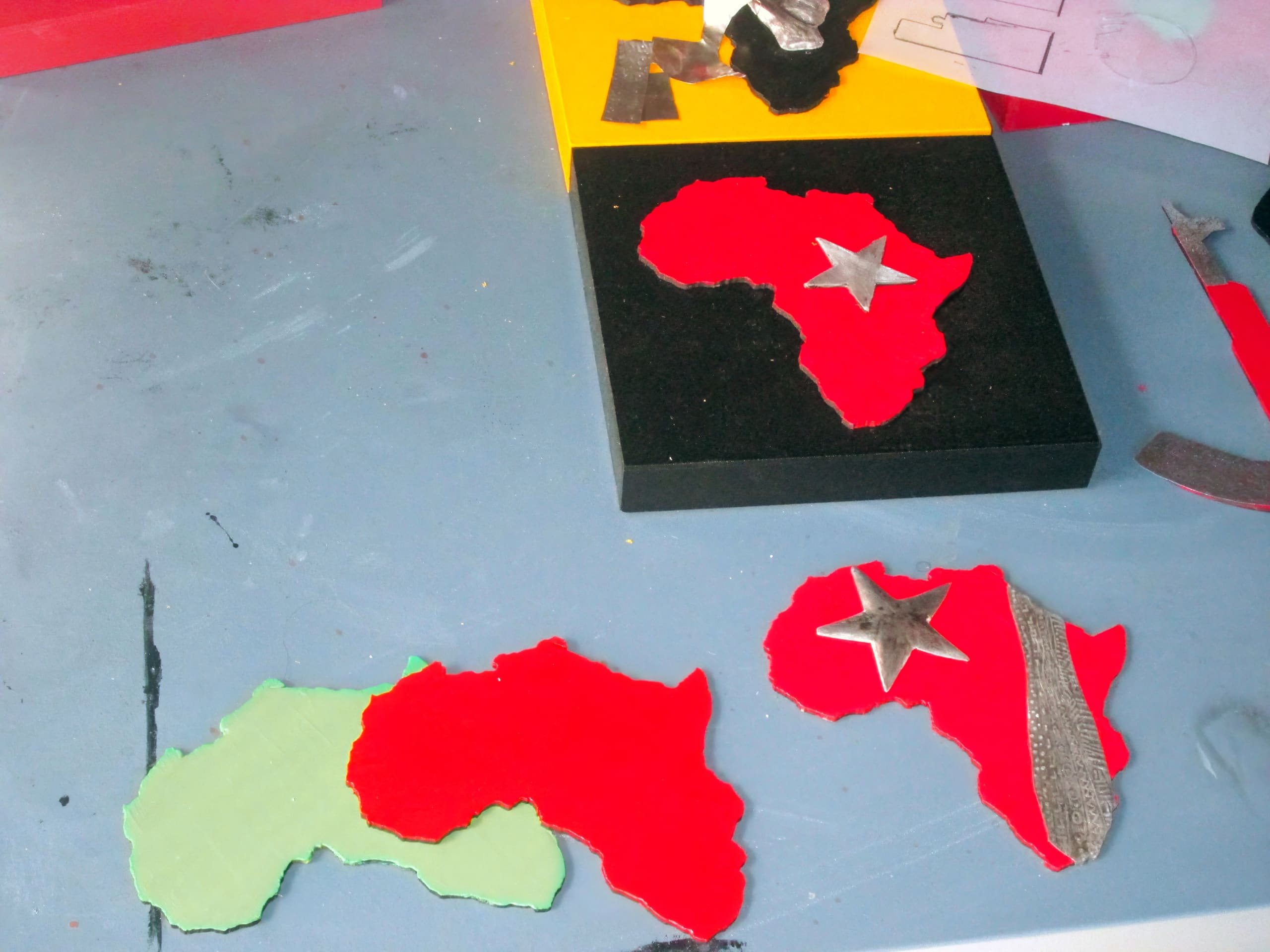 Three Africa-shaped cut-outs rest on a blue table—one green, two red. One red cut-out has a silver star and stripe. A yellow and black board displays another red Africa cut-out with a silver star. Craft materials and tools are scattered nearby.