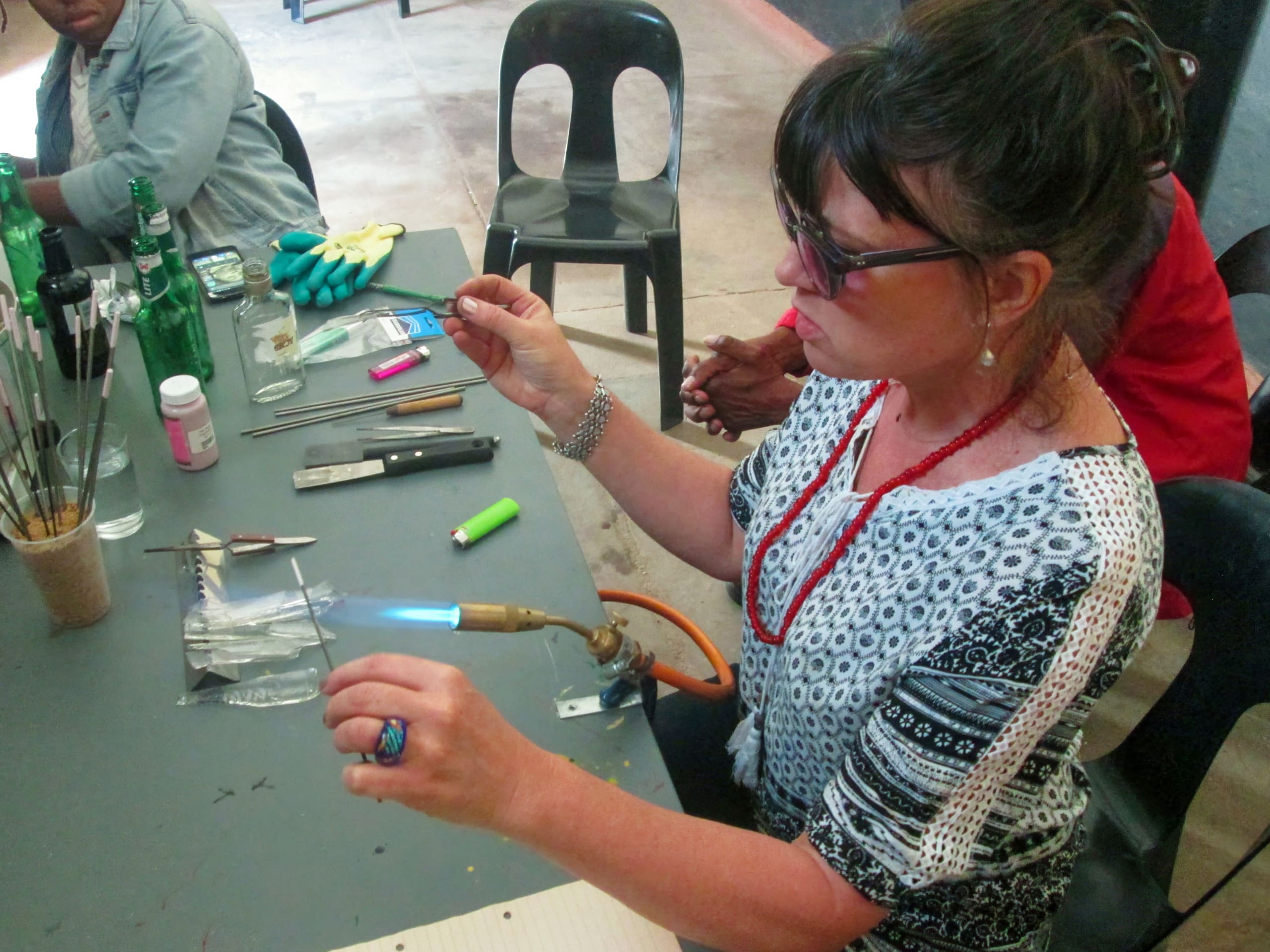 A woman wearing safety goggles uses a blowtorch to heat a small metal rod at a workbench covered with tools, glass bottles, gloves, and other materials. She focuses intently on her work, with others nearby.