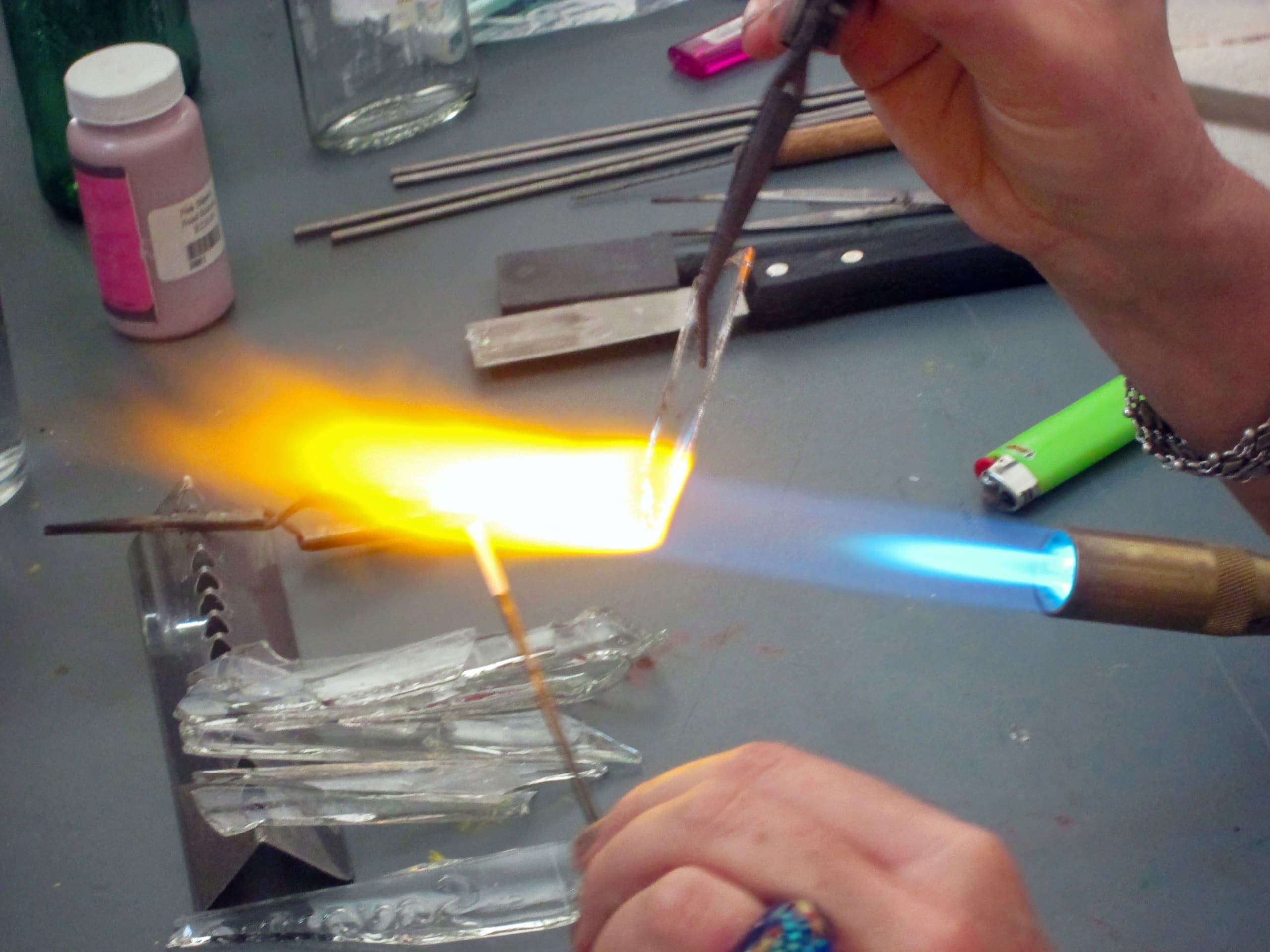 A person uses a handheld blowtorch to heat and shape clear glass rods, producing a bright orange-yellow flame. Various glass fragments, metal tools, a lighter, and jars are scattered on the work surface. Only the person's hands are visible.