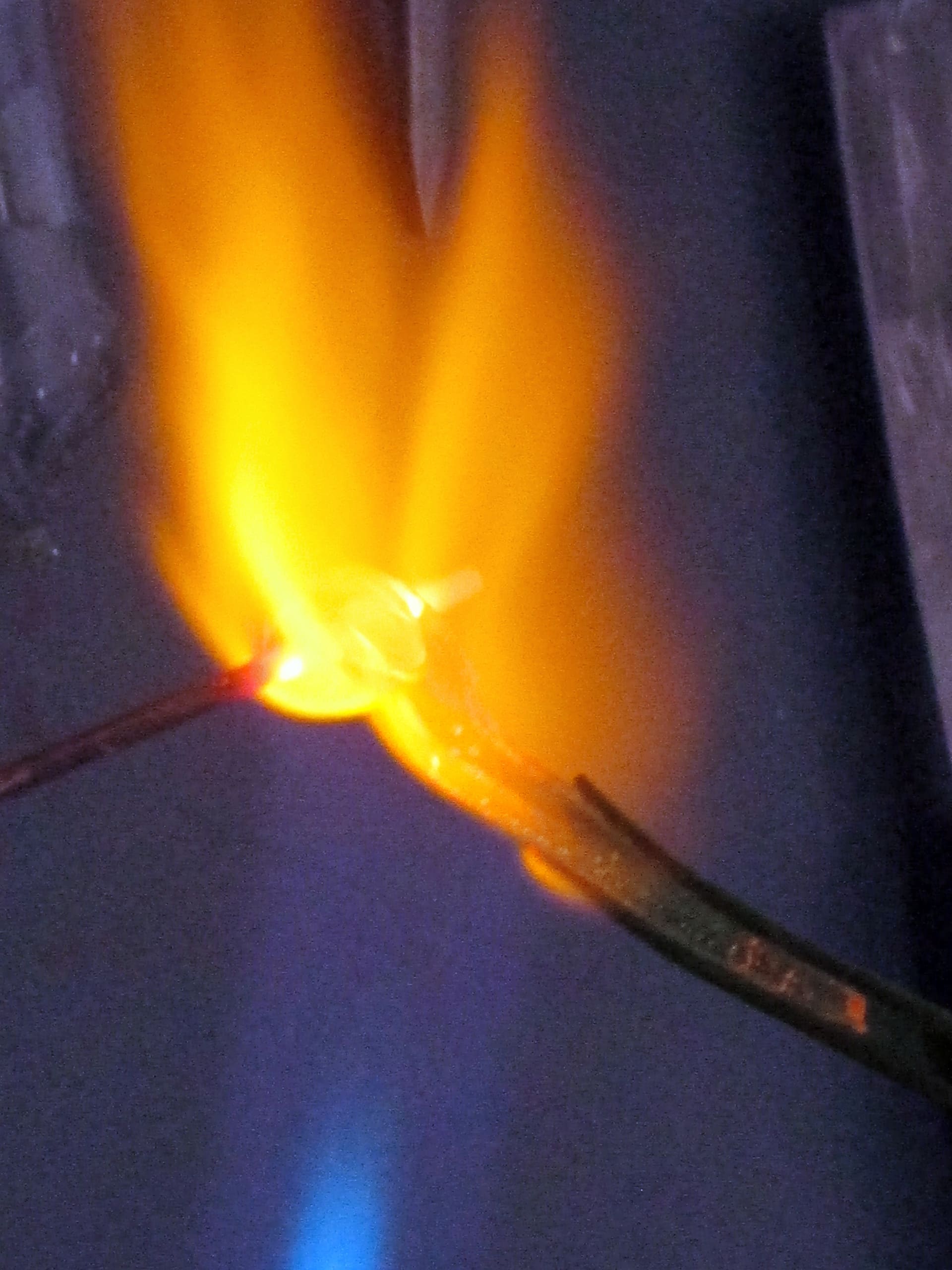 A close-up of a wire burning with bright yellow and orange flames against a dark background, with glowing embers visible at the heated area where the fire is most intense.