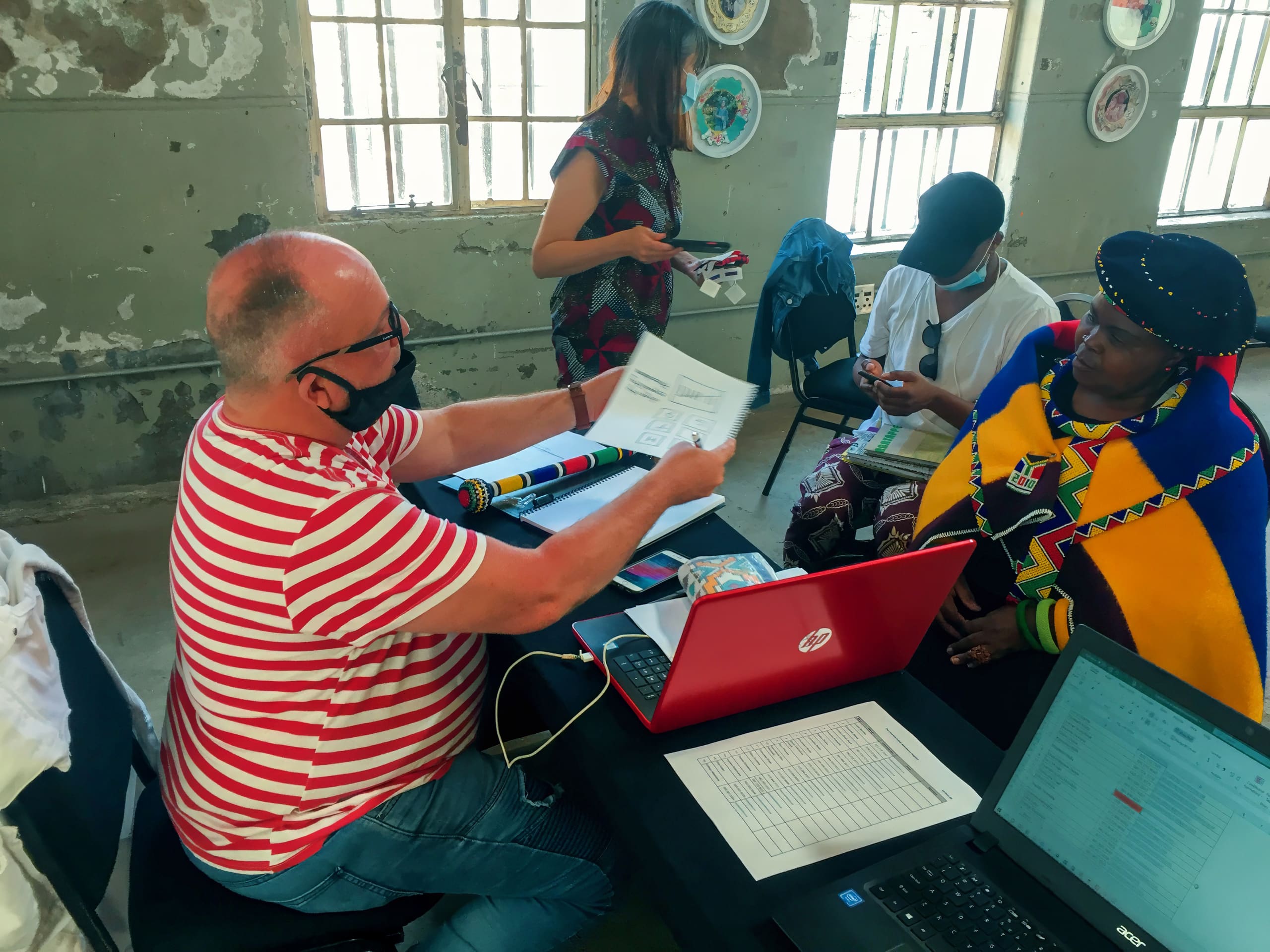 A man in a red striped shirt and black mask hands papers to a woman in colourful traditional attire. They sit at a table with two laptops and documents. Two other people, one wearing a mask, are in the background inside a sunlit, worn room.