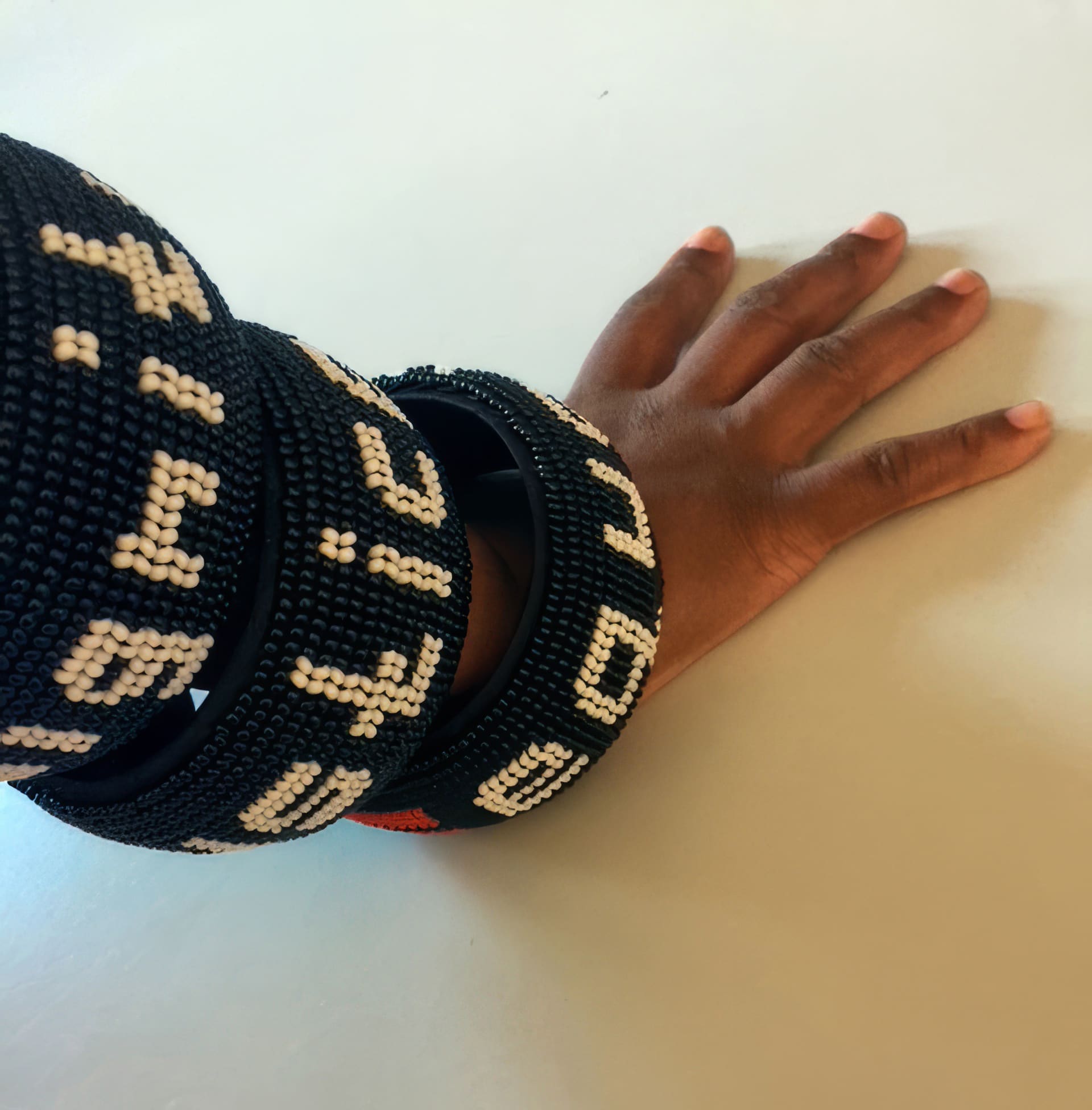 A hand with medium-brown skin rests on a white surface, wearing three large, black beaded bracelets with white geometric patterns and text-like symbols. The bracelets appear chunky and are stacked on the wrist and forearm.