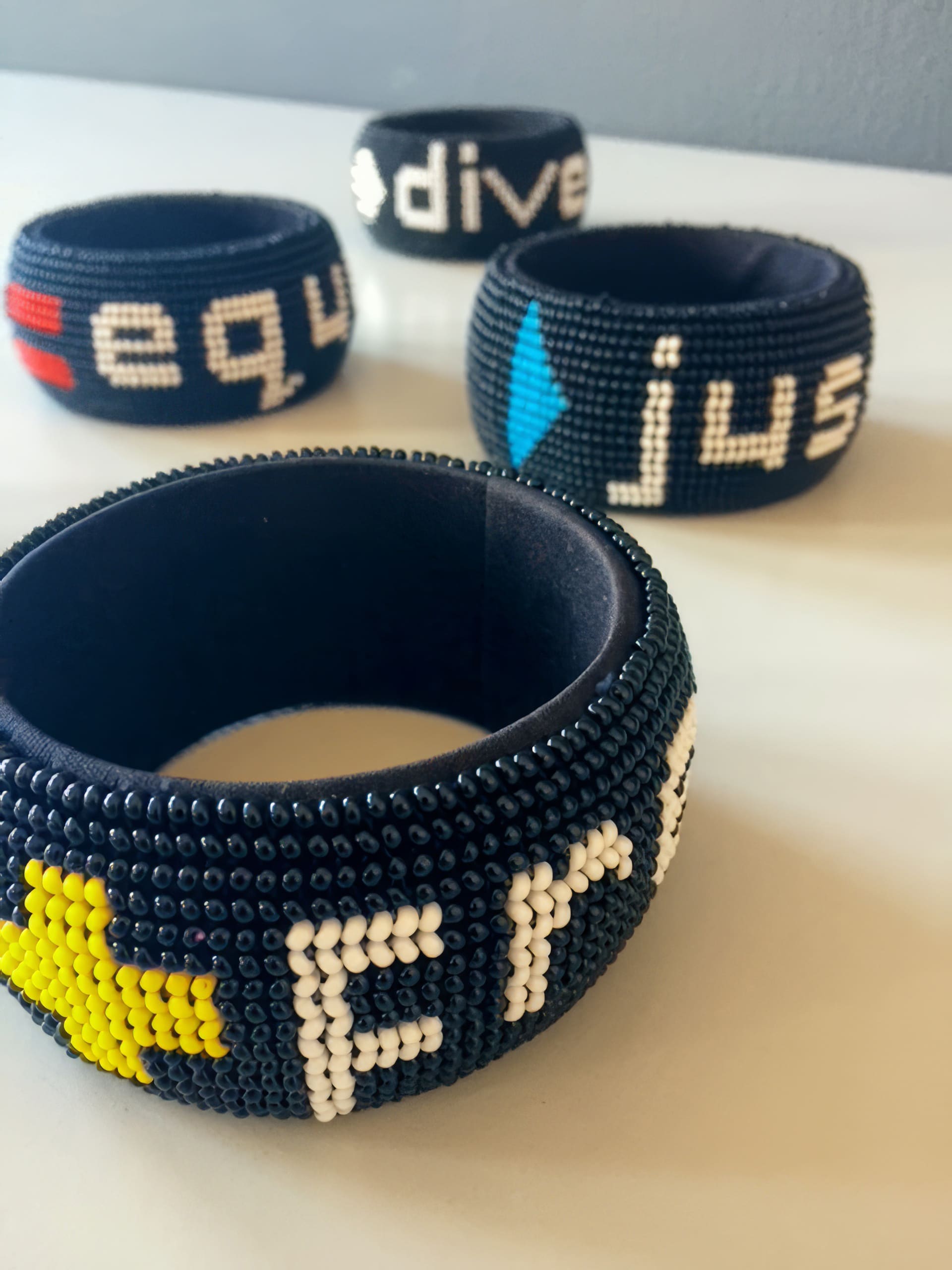 Four black beaded bracelets rest on a white surface. Each bracelet features colourful beadwork spelling out positive words, such as “free,” “just,” “equ,” and “div”, along with small star or arrow shapes in yellow, blue, red, and white.
