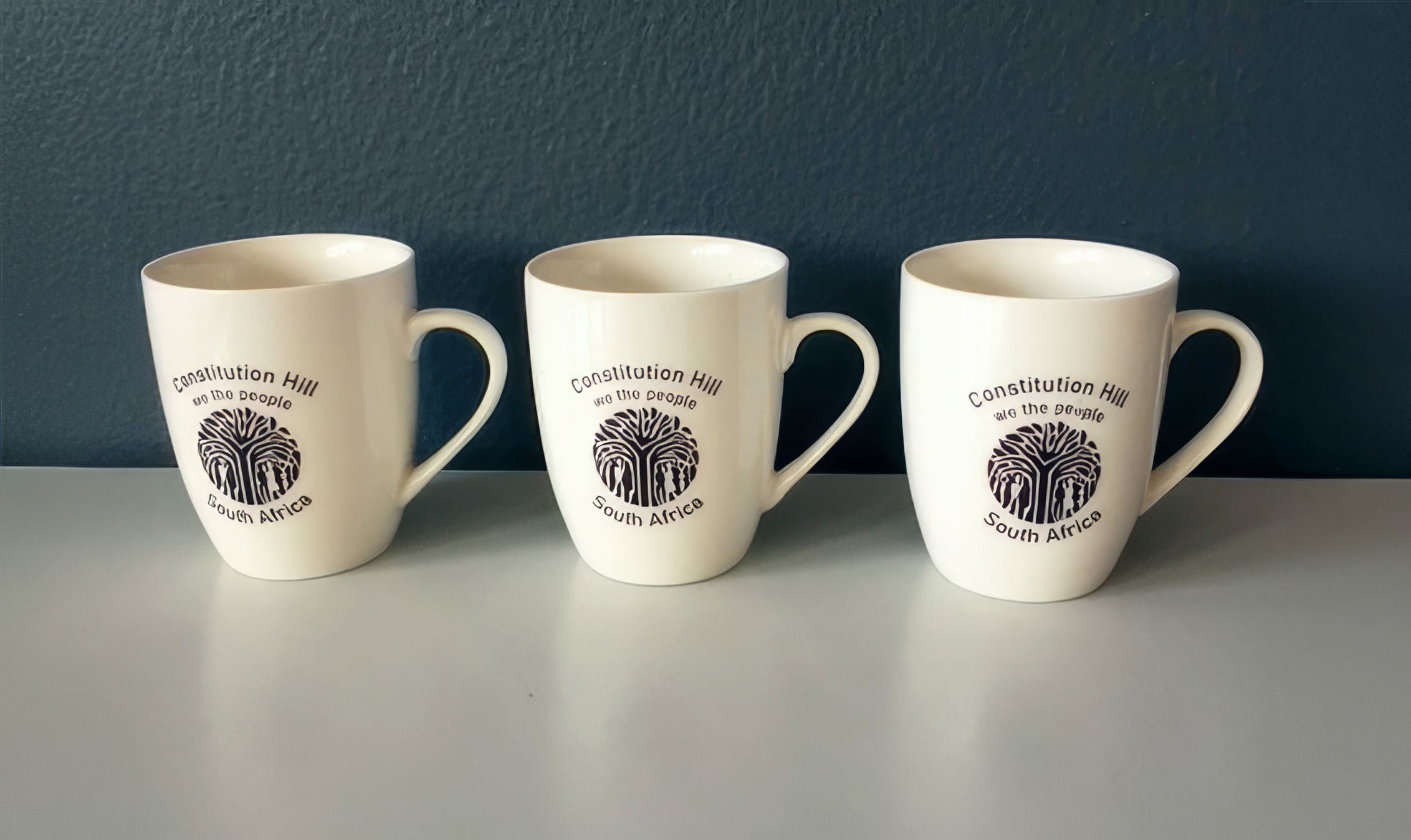 Three white ceramic mugs sit side by side on a light grey surface against a dark blue wall. Each mug has a handle and features the same black logo and text: “Constitution Hill, We the People, South Africa” with a stylised design below the text.