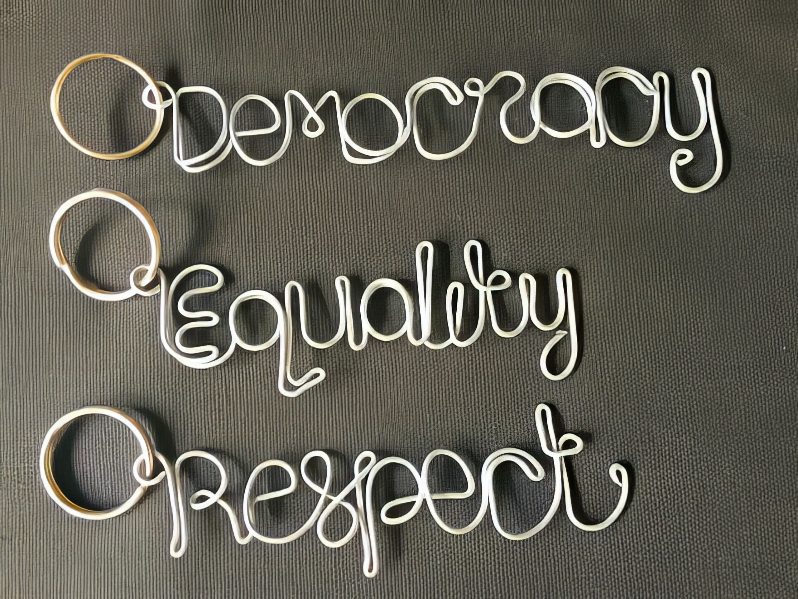 Three keyrings shaped from wire spell out “Democracy,” “Equality,” and “Respect” in cursive writing. The words are arranged one below the other on a textured dark background. The wire is light-coloured, standing out against the surface.