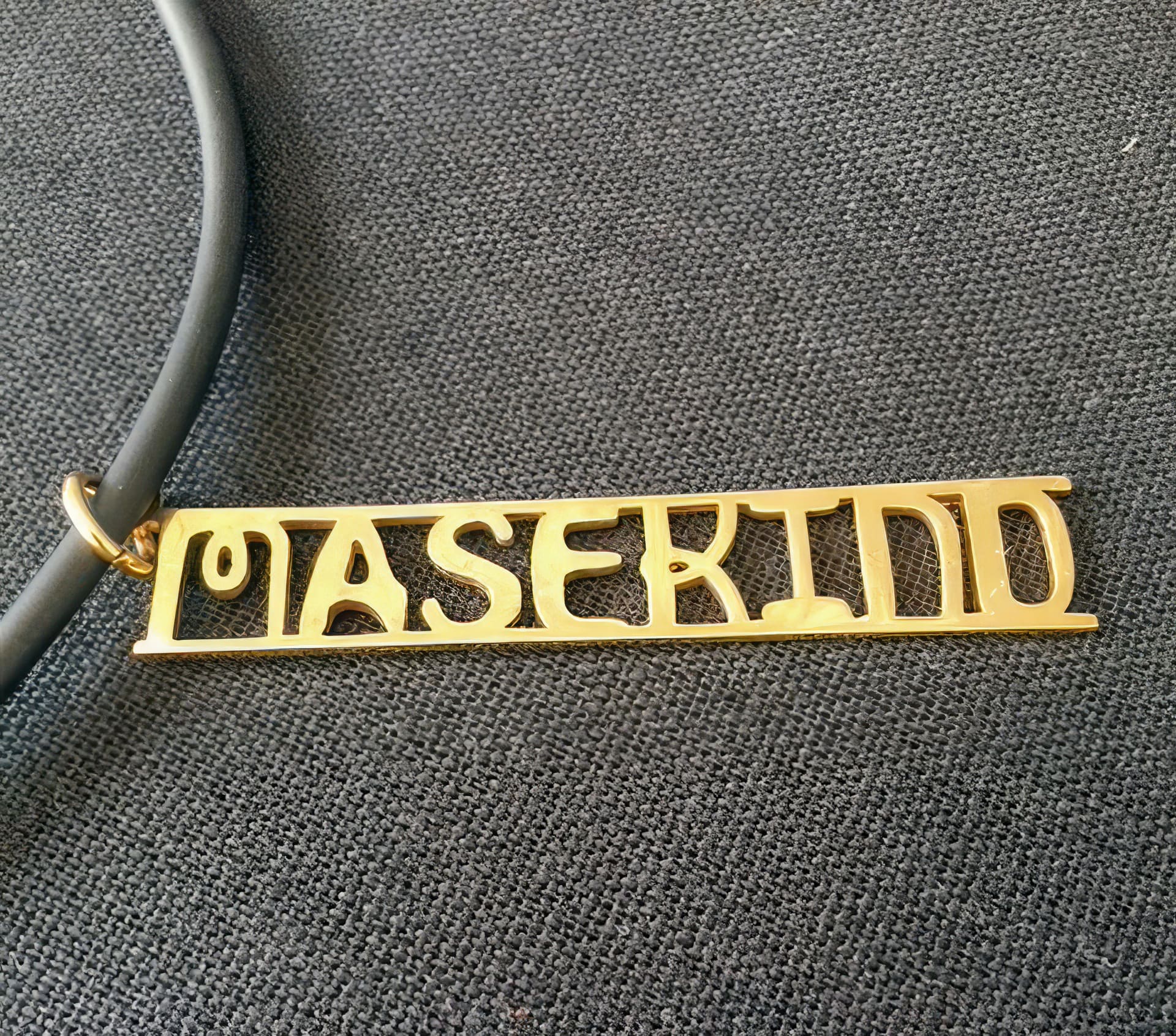 A gold rectangular pendant with the word “MASKIND” in stylised cut-out letters hangs from a black cord, resting on a textured dark grey fabric background. The pendant has smooth, shiny edges and an attached gold loop for the cord.