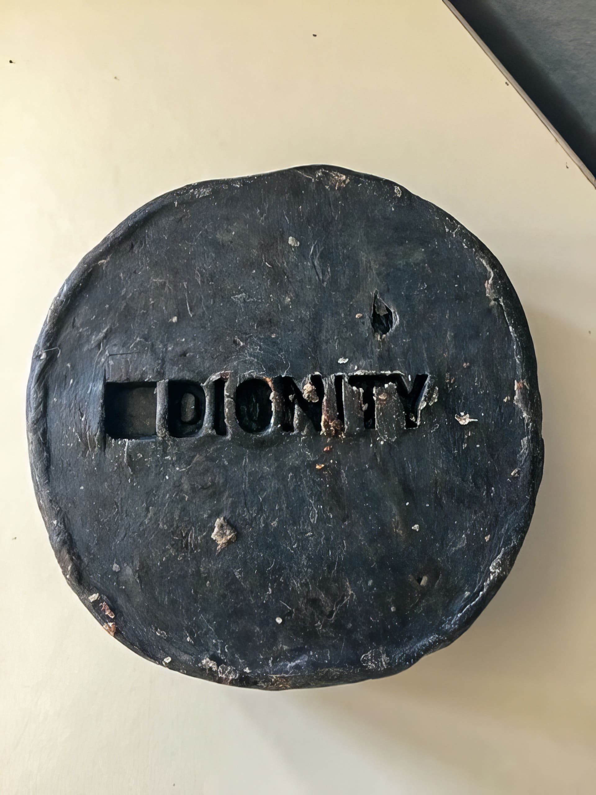 A round, dark, textured object, possibly clay or metal, rests on a light surface. The word 