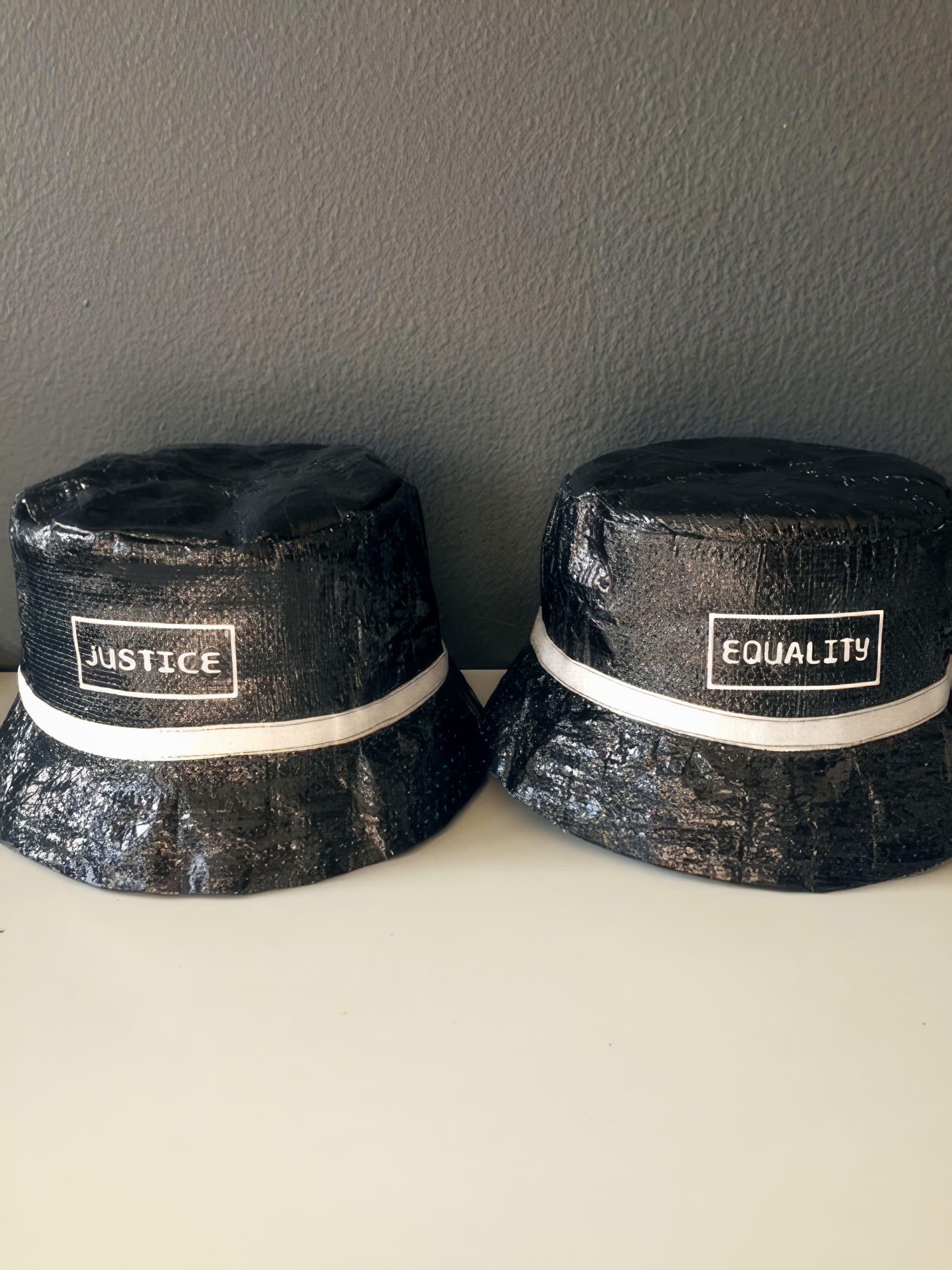 Two black bucket hats sit side by side on a white surface, each featuring a white ribbon and a rectangular white patch. The left hat reads 