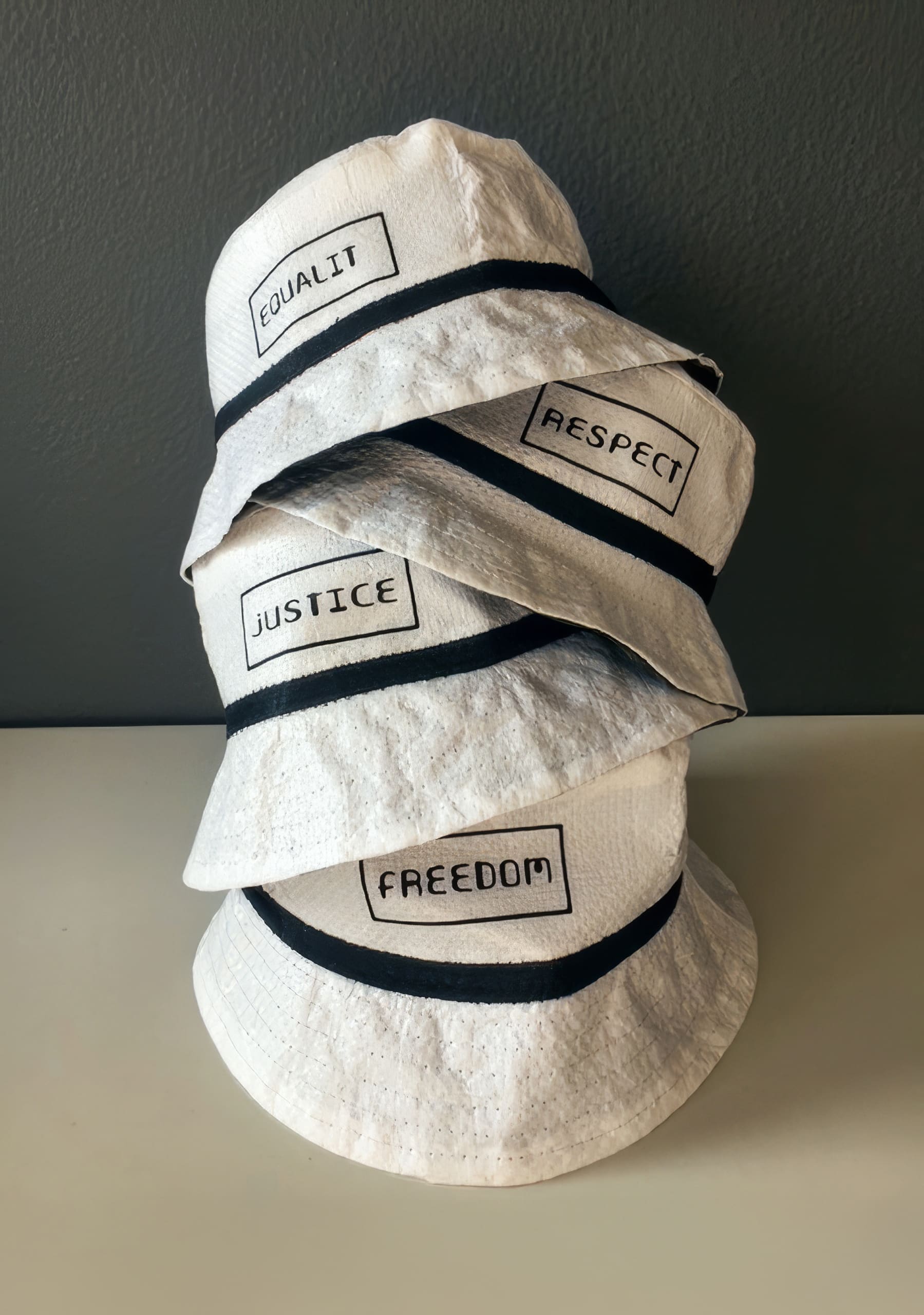 Four white bucket hats are stacked on a white surface against a dark wall. Each hat has a black band and a rectangular patch with black text: 