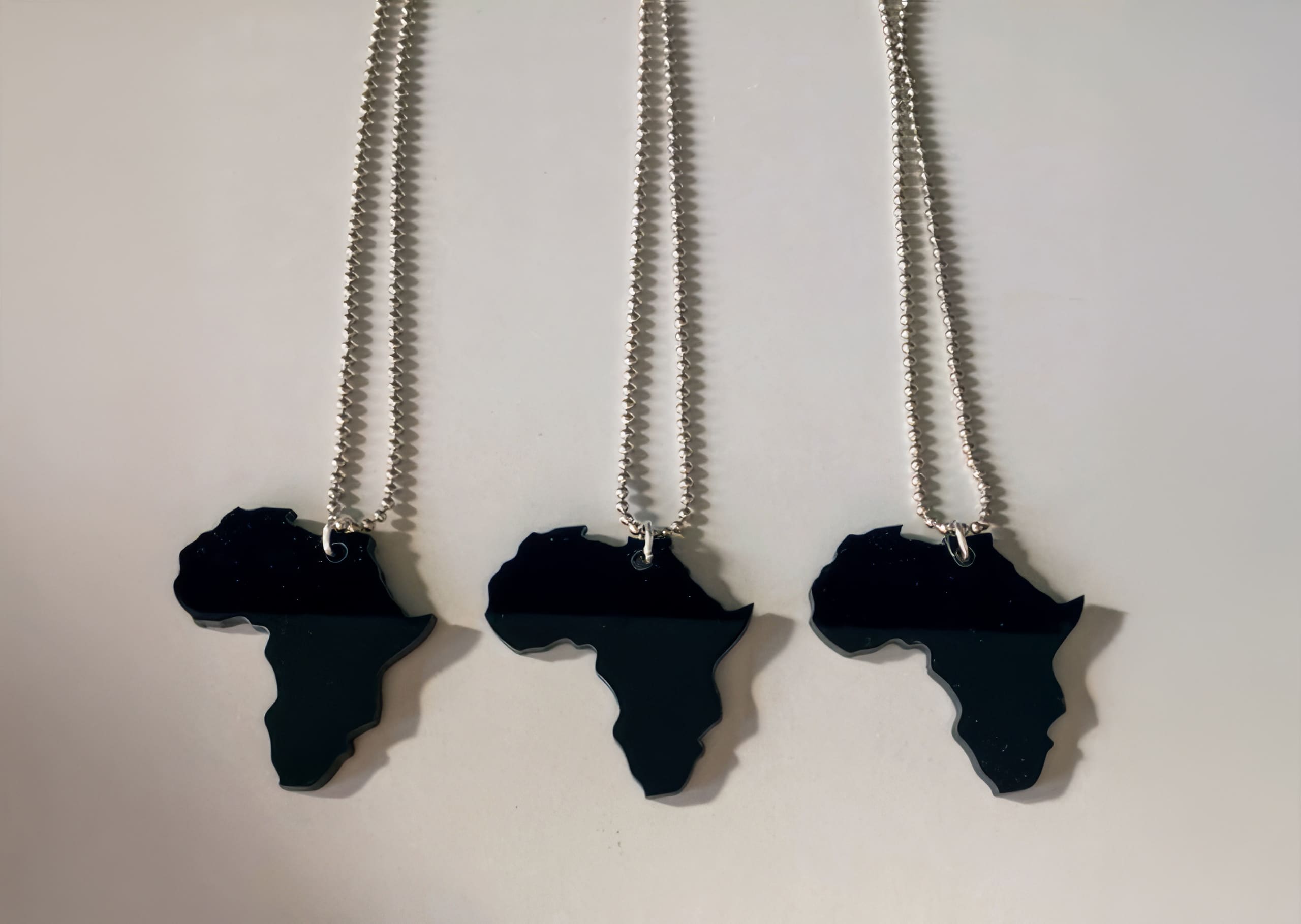 Three silver chains with black pendants shaped like the African continent are laid out vertically on a white surface, evenly spaced and parallel to each other. The pendants are glossy and reflect light, highlighting their distinct Africa silhouette.