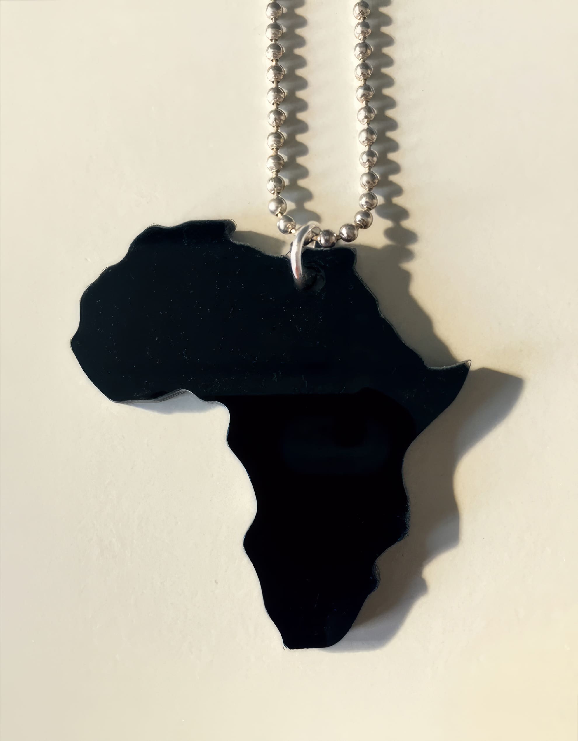 A black pendant shaped like the African continent hangs from a silver beaded chain. The pendant casts a shadow on a light, cream-coloured surface, creating a contrast between the dark shape and the pale background.