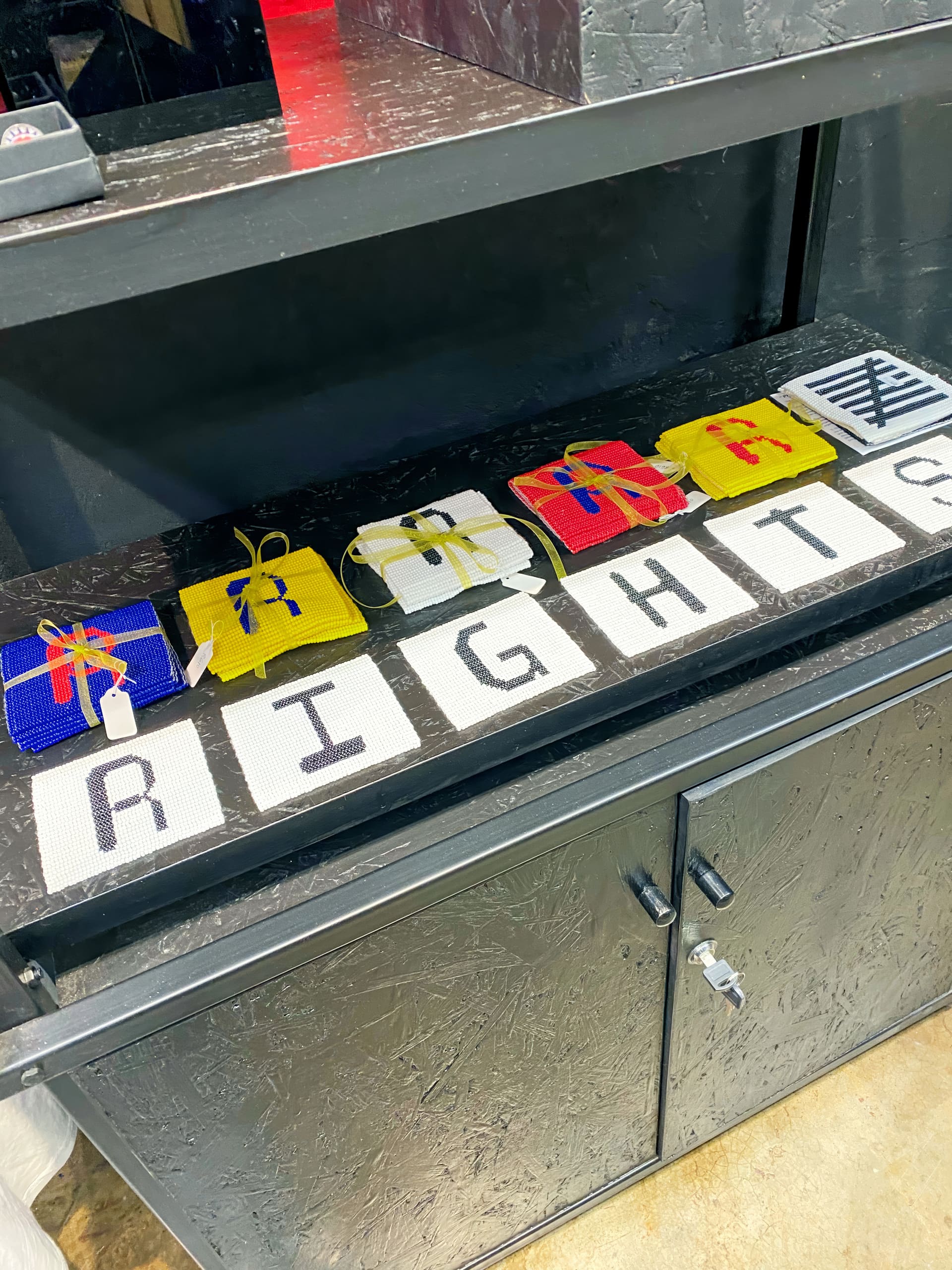 A display shelf holds six colourful woven pouches tied with string, each above a white square with a black letter. Together, the squares spell “RIGHTS.” The shelf and background are black, giving a bold contrast to the vibrant pouches and letters.