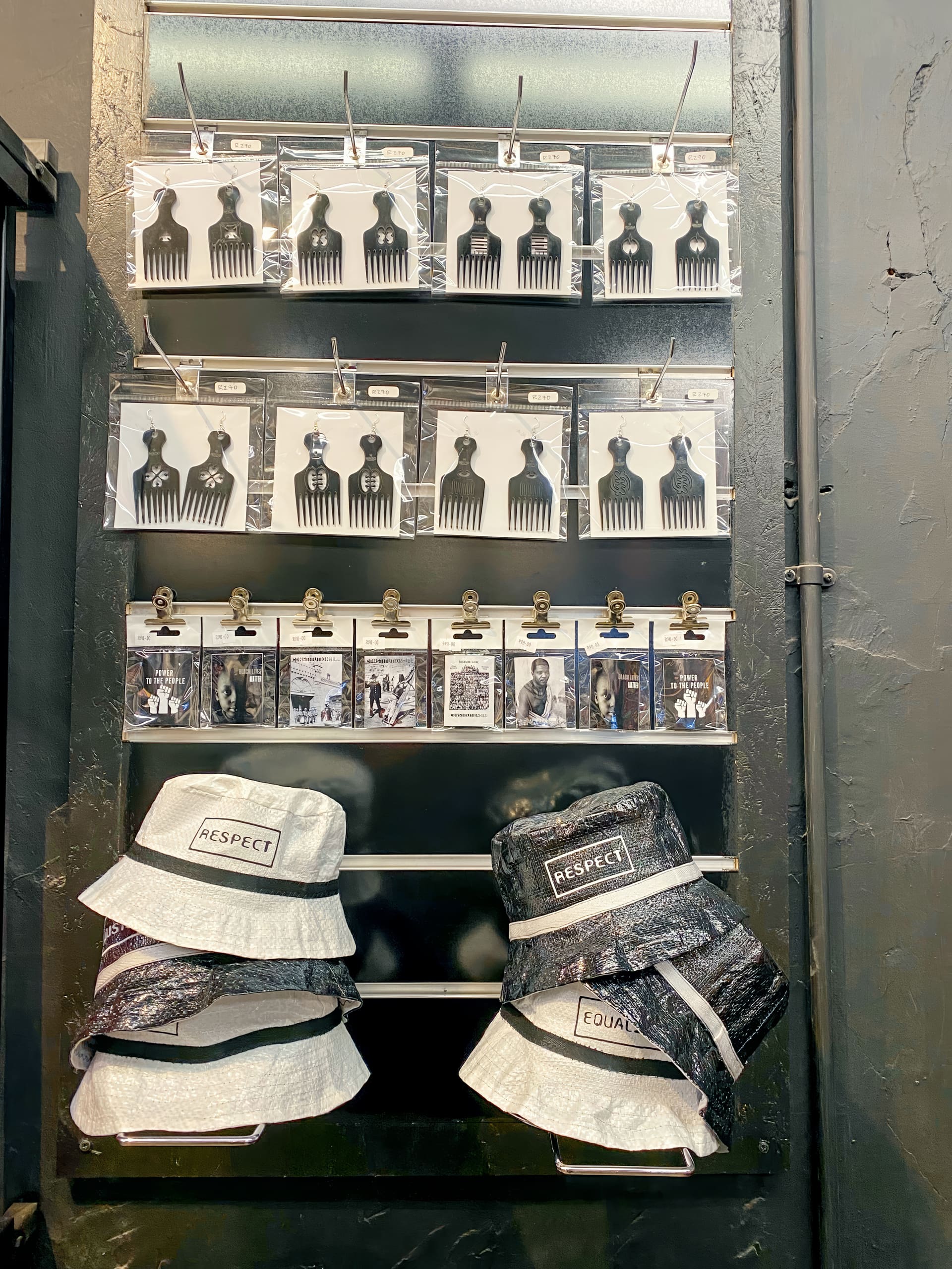 A black display wall features metal afro combs and black-and-white prints of people attached with clips. Two hats labelled 