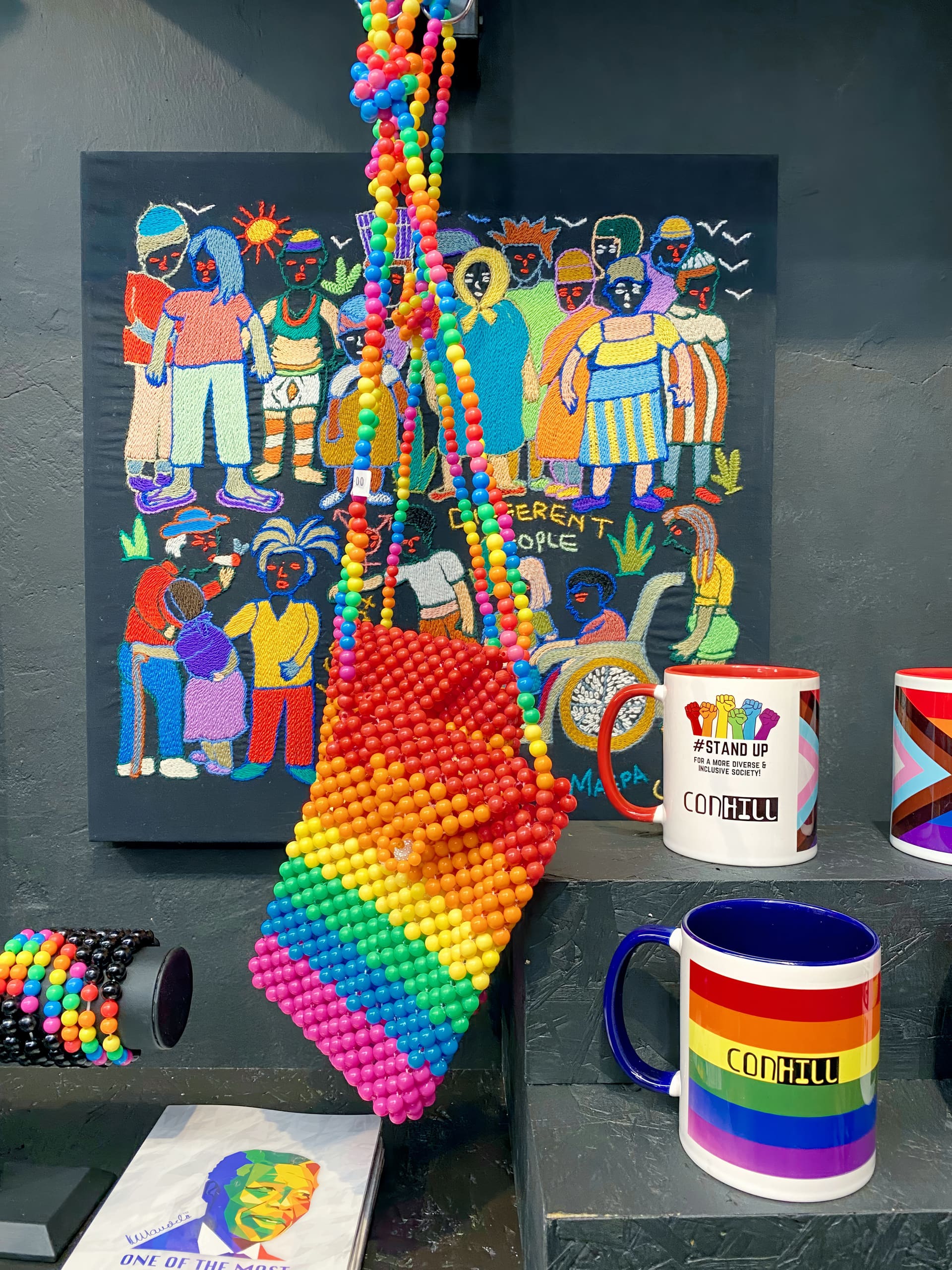 A bright beaded rainbow bag hangs in front of a colourful painting of diverse LGBTQ+ people. Nearby are rainbow mugs, one with “CONHILL” and another with “#STAND UP FOR EQUALITY.” A rainbow beaded bracelet and rainbow-patterned items surround them.