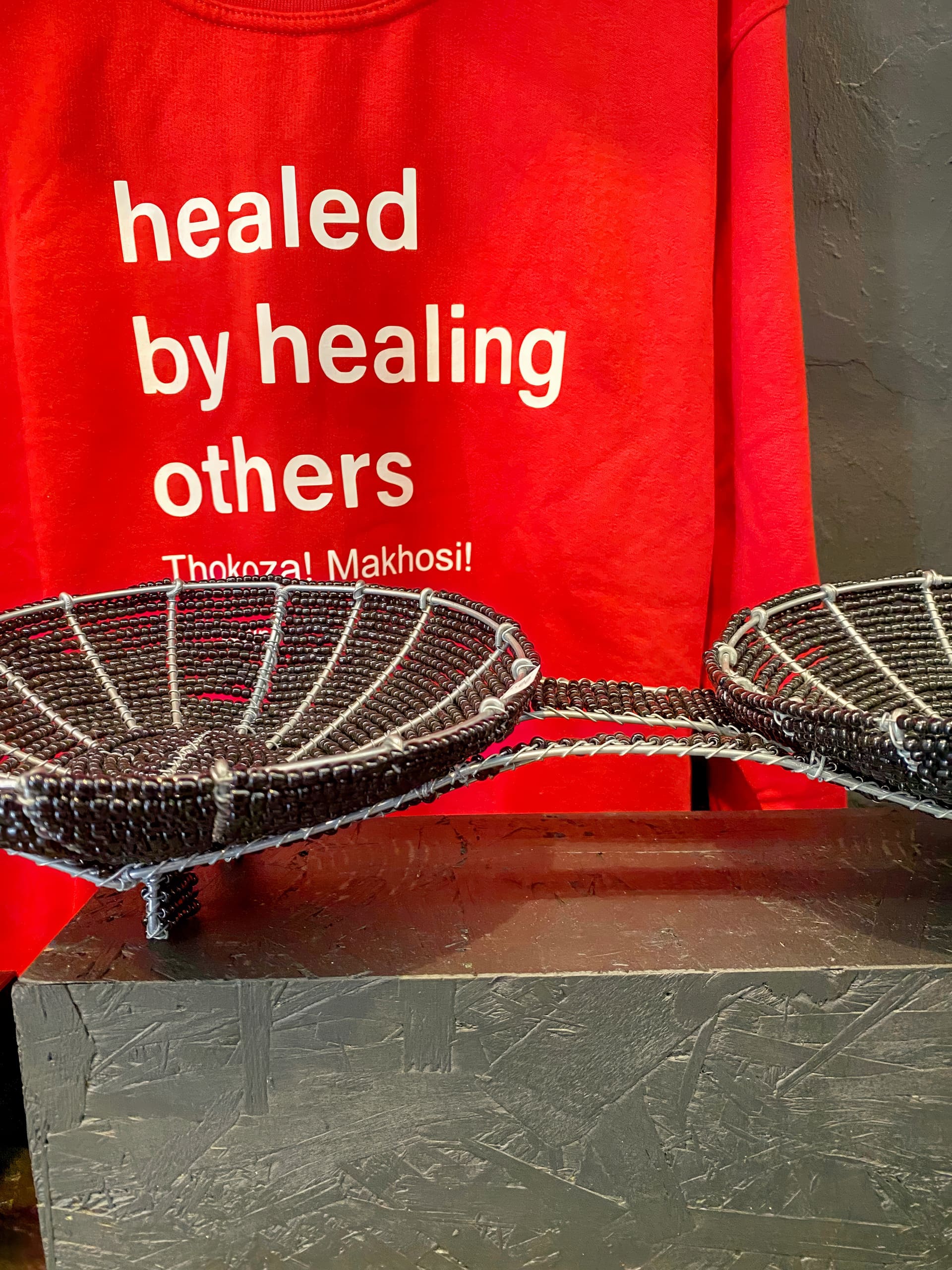 A red shirt with white text reading “healed by healing others Thokoza! Makhosi!” hangs behind two black, round, woven tray-like baskets displayed on a dark, textured surface. The background wall is grey and partially visible.