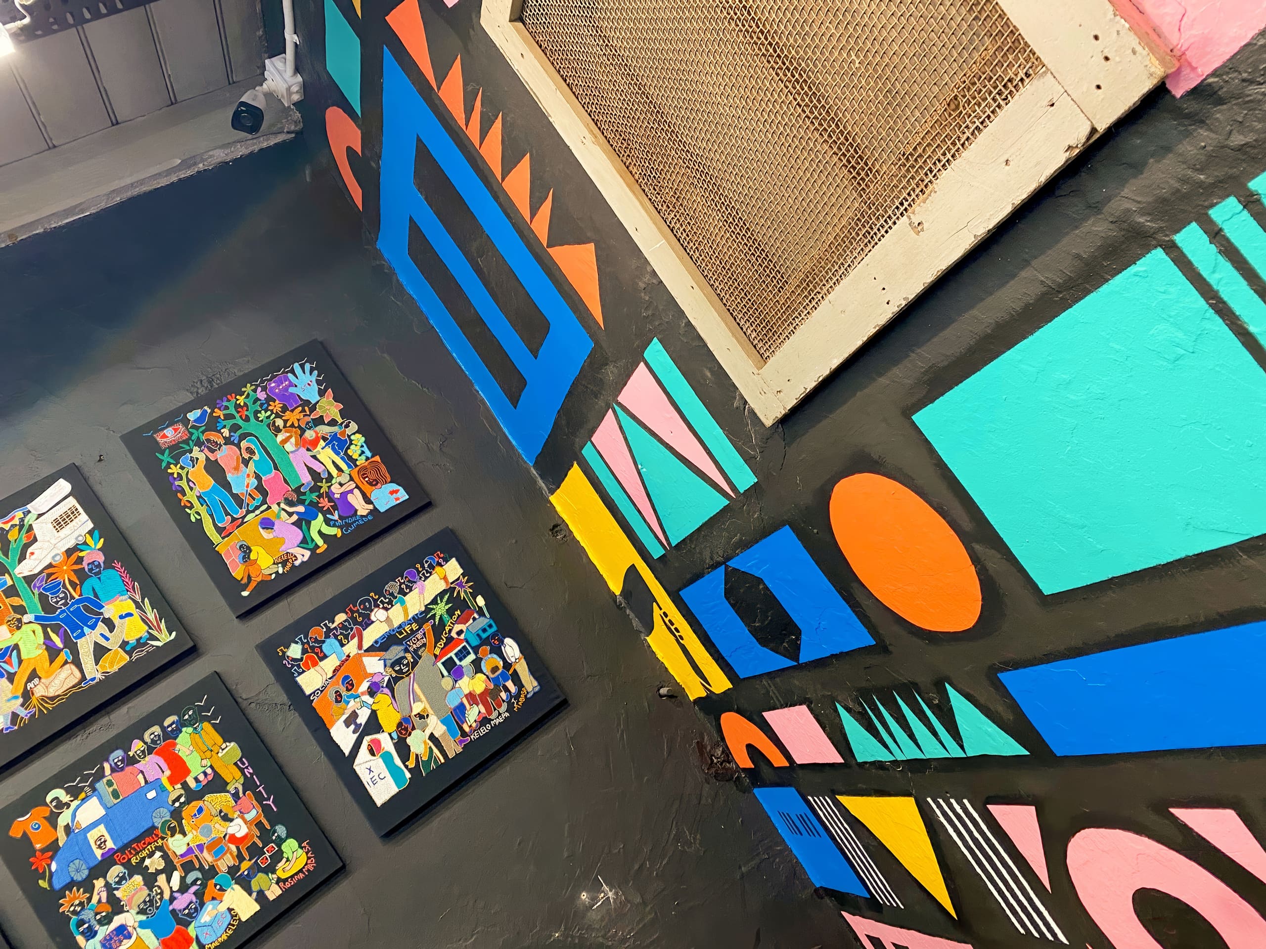 Colourful geometric murals with bold shapes (triangles, circles, rectangles) are painted on black walls. Three framed, vibrant, cartoon-style artworks hang nearby. Part of a metal mesh vent appears above, adding texture to the eclectic scene.