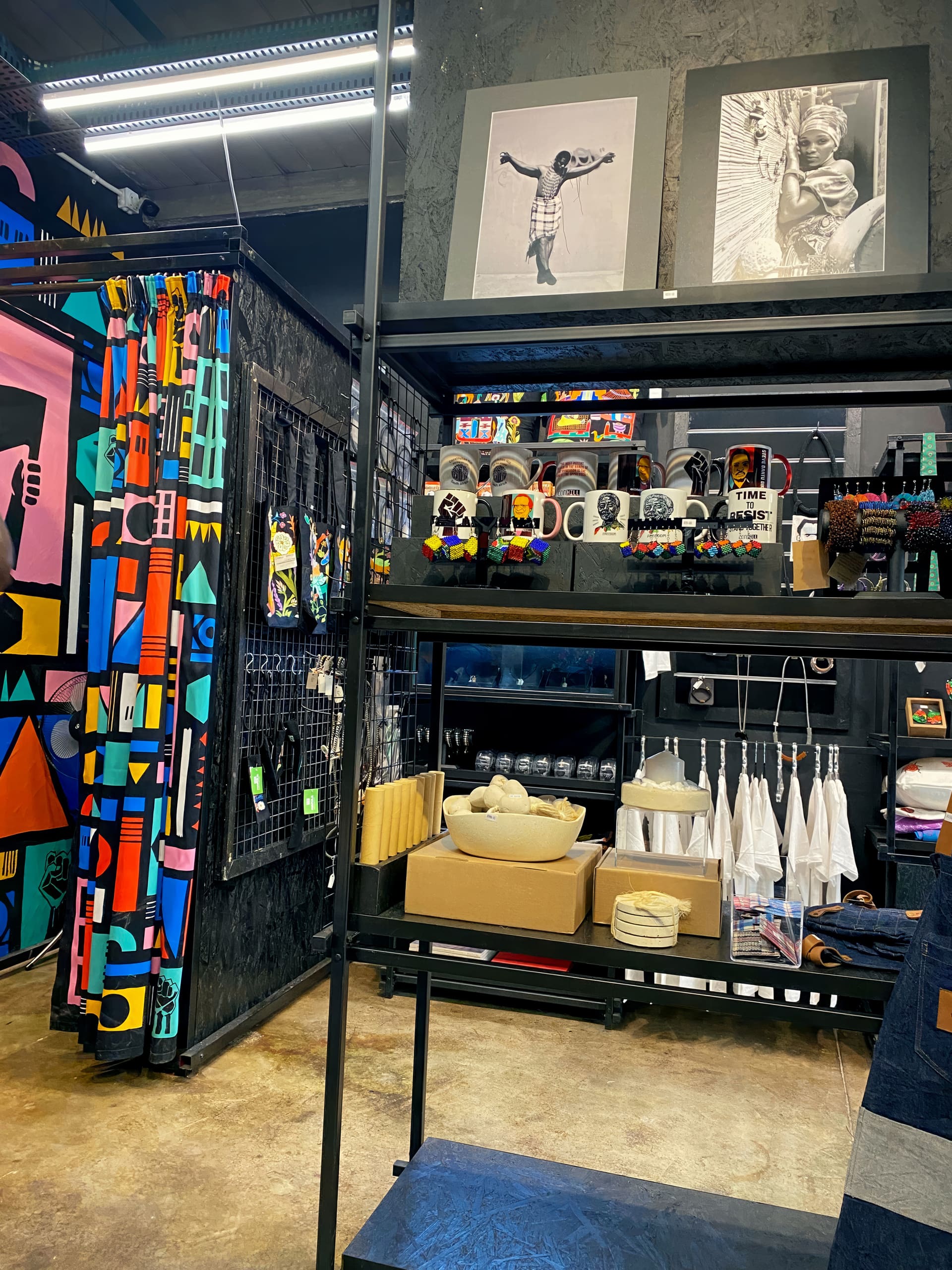 A colourful shop display features vibrant patterned fabrics, art prints of children on a grey wall, mugs with bold designs, beaded jewellery, white aprons, stacked boxes, and shelves with folded textiles. The store has an artistic, modern, and eclectic style.