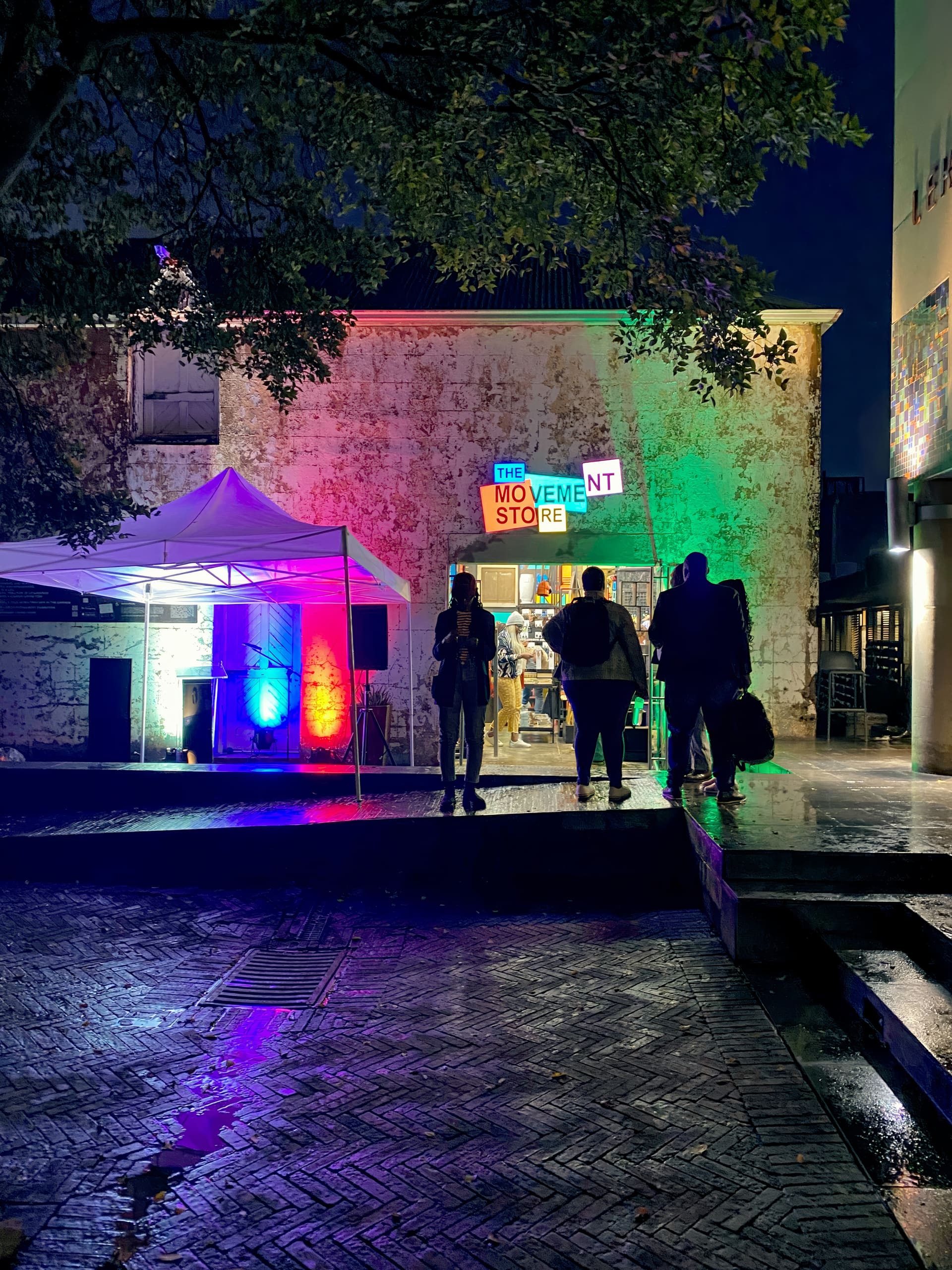 People stand in front of The Movement Shop at night, illuminated by colourful lights (red, green, blue). A white marquee and speakers are set up nearby. Wet pavement and leafy tree branches frame the lively, urban outdoor scene.