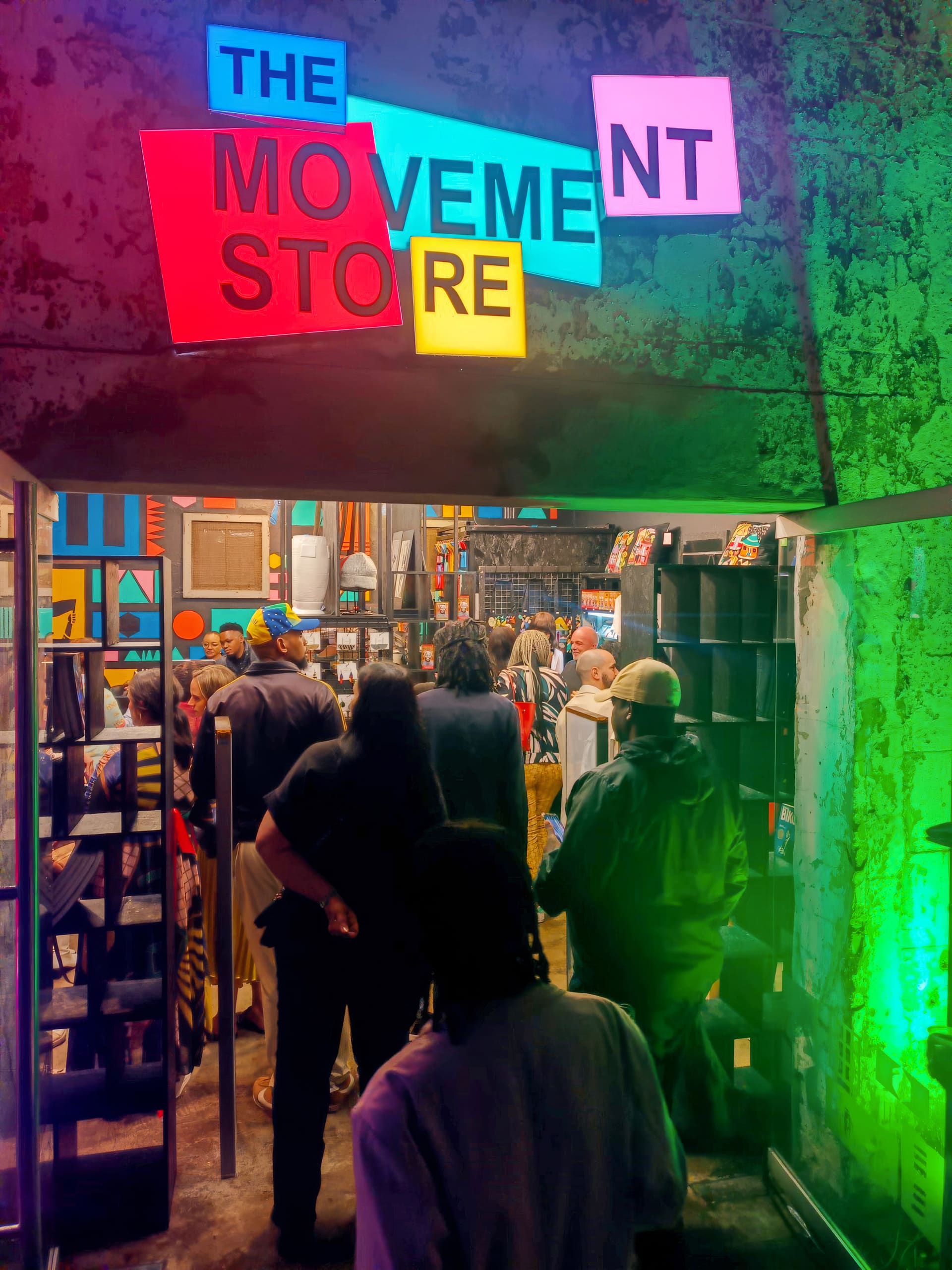 A group of people gathers inside a colourful, well-lit shop filled with shelves and art. A large, bright sign above reads “THE MOVEMENT SHOP” in multicoloured block letters. The walls and displays feature vivid decor and creative arrangements.