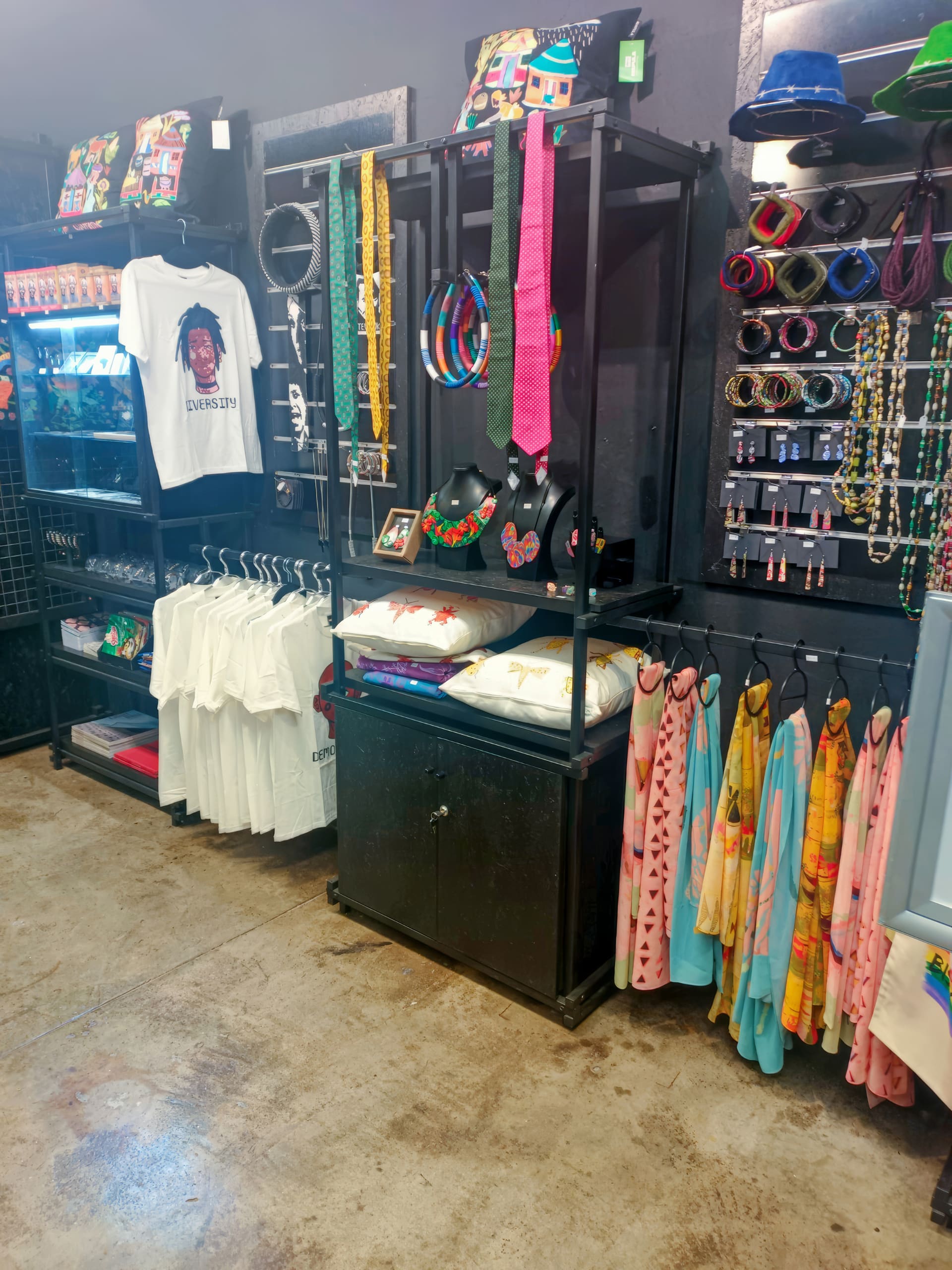 A boutique display with colourful jewellery, hats, and patterned wristbands on black shelves. Hanging below are bright scarves, white T-shirts with 