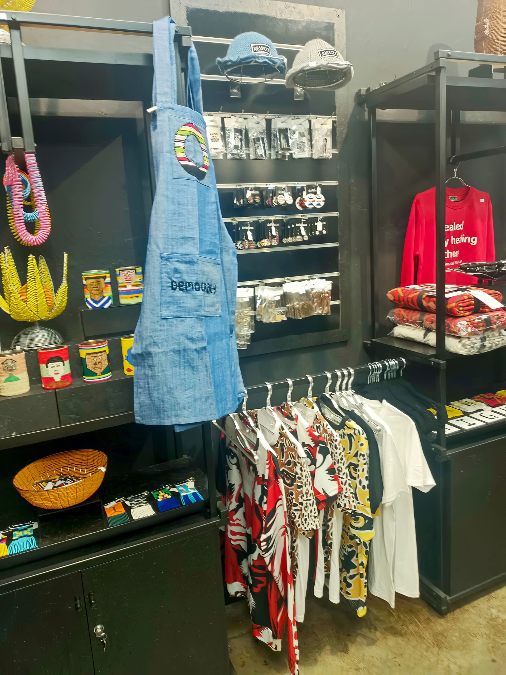 A shop display features colourful mugs, beaded décor, a blue apron with embroidery, folded red shirts with white text, patterned T-shirts on hangers, hats, and small souvenirs on black shelves against a grey wall.