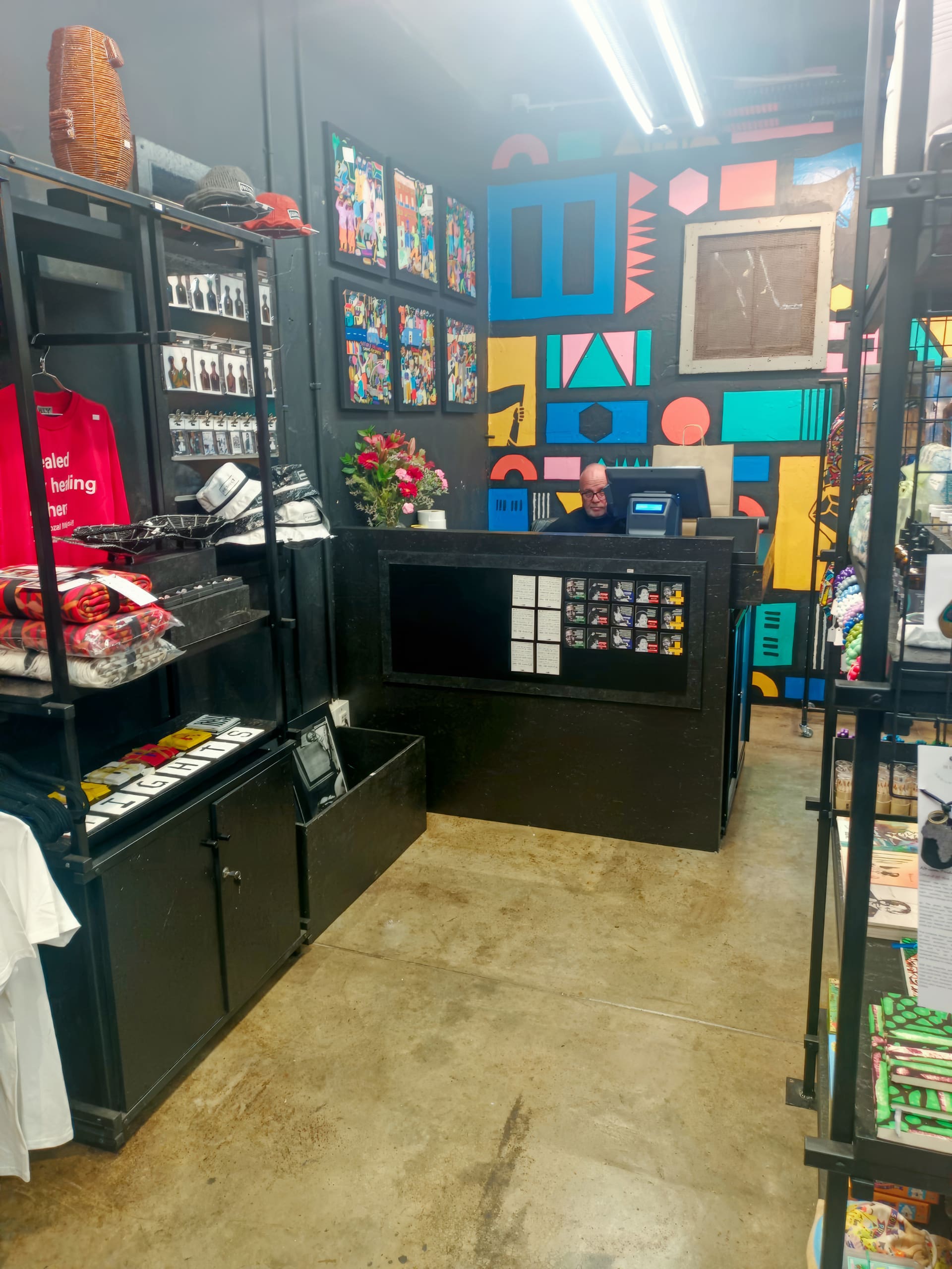 A small, colourful shop interior with a person sitting behind a black counter. Bright geometric art covers the back wall. Shelves display clothing, hats, accessories, and framed pictures. The concrete floor and black shelves contrast with the vivid decor.
