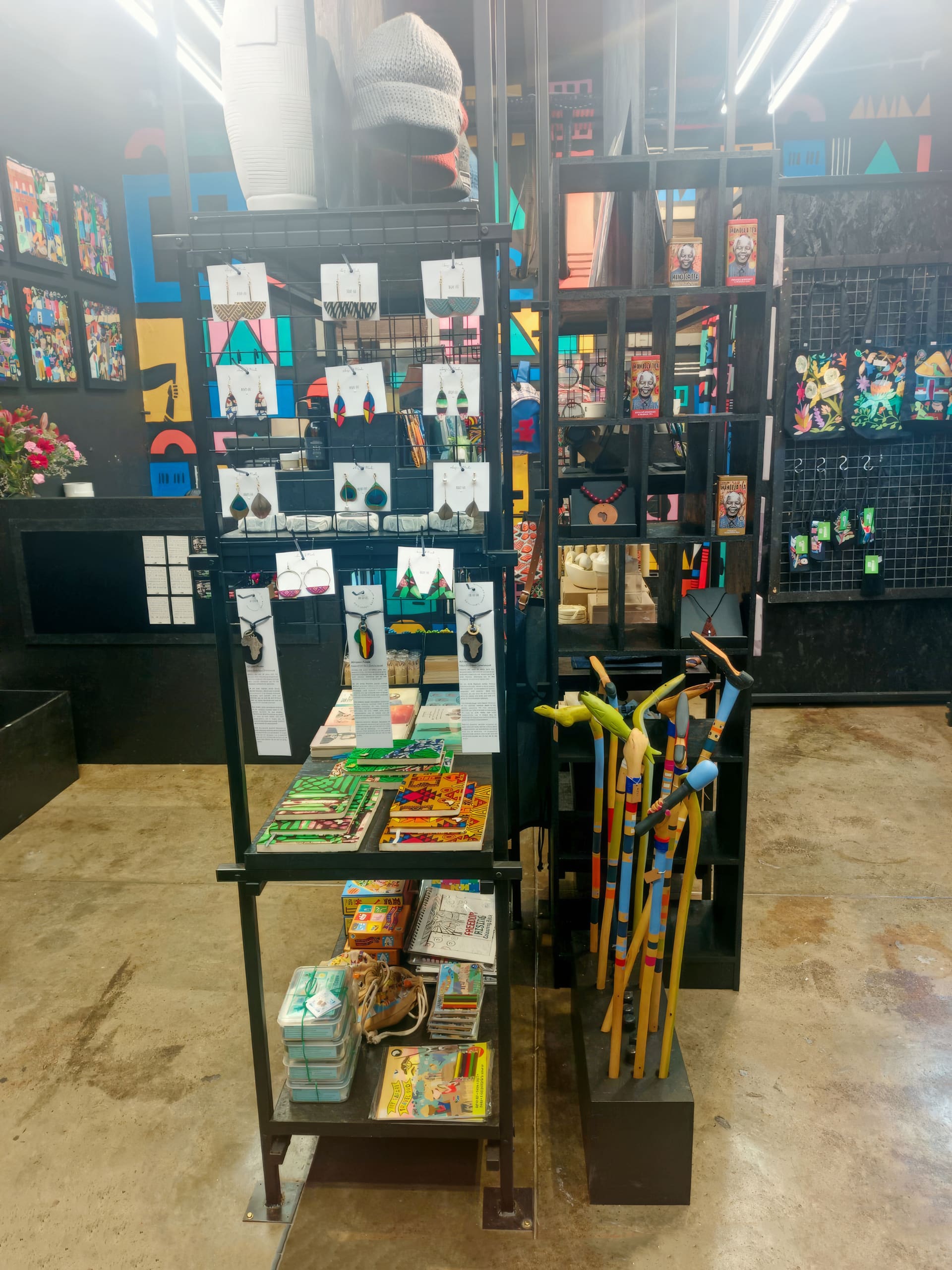 A shop display showcases colourful walking sticks, art supplies, jewellery, and accessories. Framed vibrant artwork covers the back wall. The floor is polished concrete, and shelves and racks are made of black metal, creating an artistic, modern retail space.