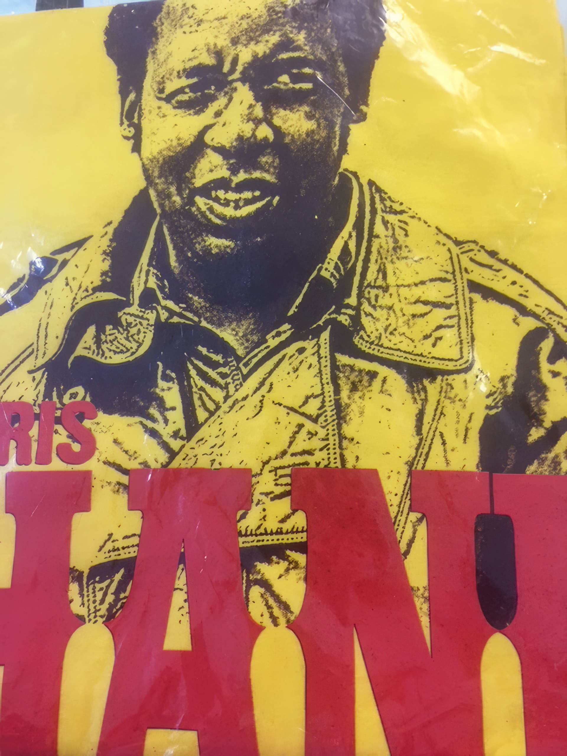 A yellow poster shows a black-and-white portrait of a person wearing a jacket, smiling slightly. Large red block letters partially obscure the lower part of the image. The design has a bold, graphic look.