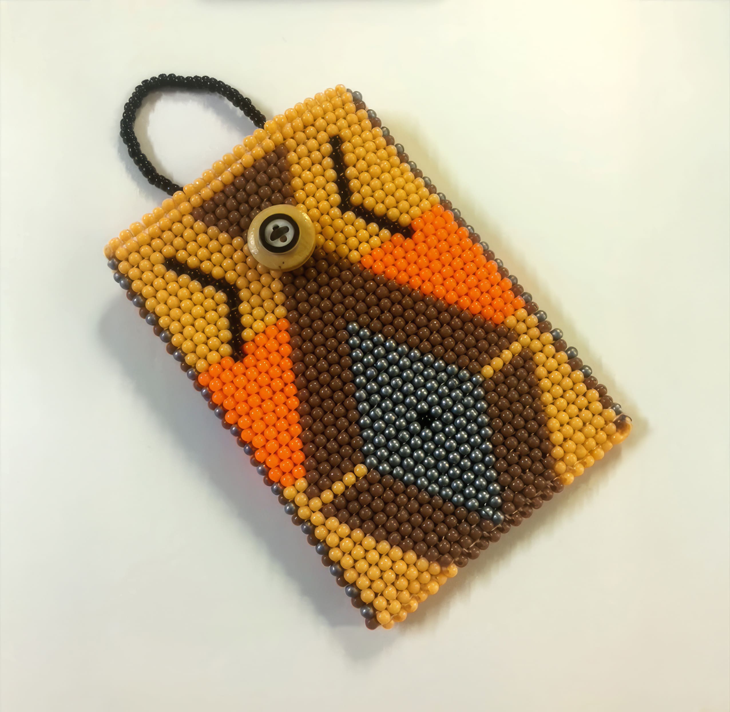 A handmade beaded pouch with a geometric pattern in shades of brown, orange, yellow, and black. It has a black beaded loop handle and a tan button for fastening, all set against a plain white background.