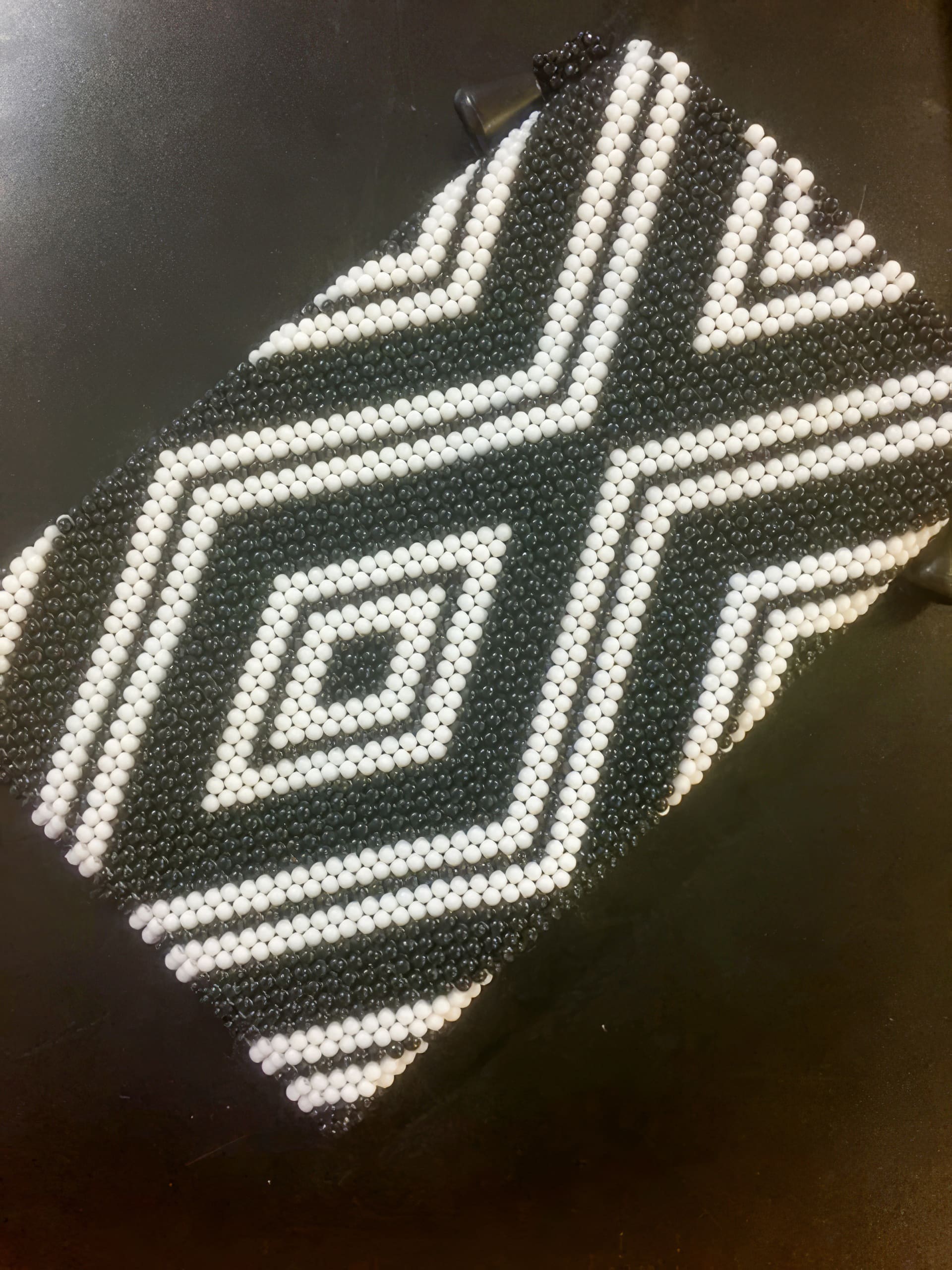 A rectangular pouch covered in black and white beads arranged in geometric diamond and zigzag patterns, photographed on a dark, flat surface. The beadwork creates contrasting shapes that form a bold, symmetrical design.