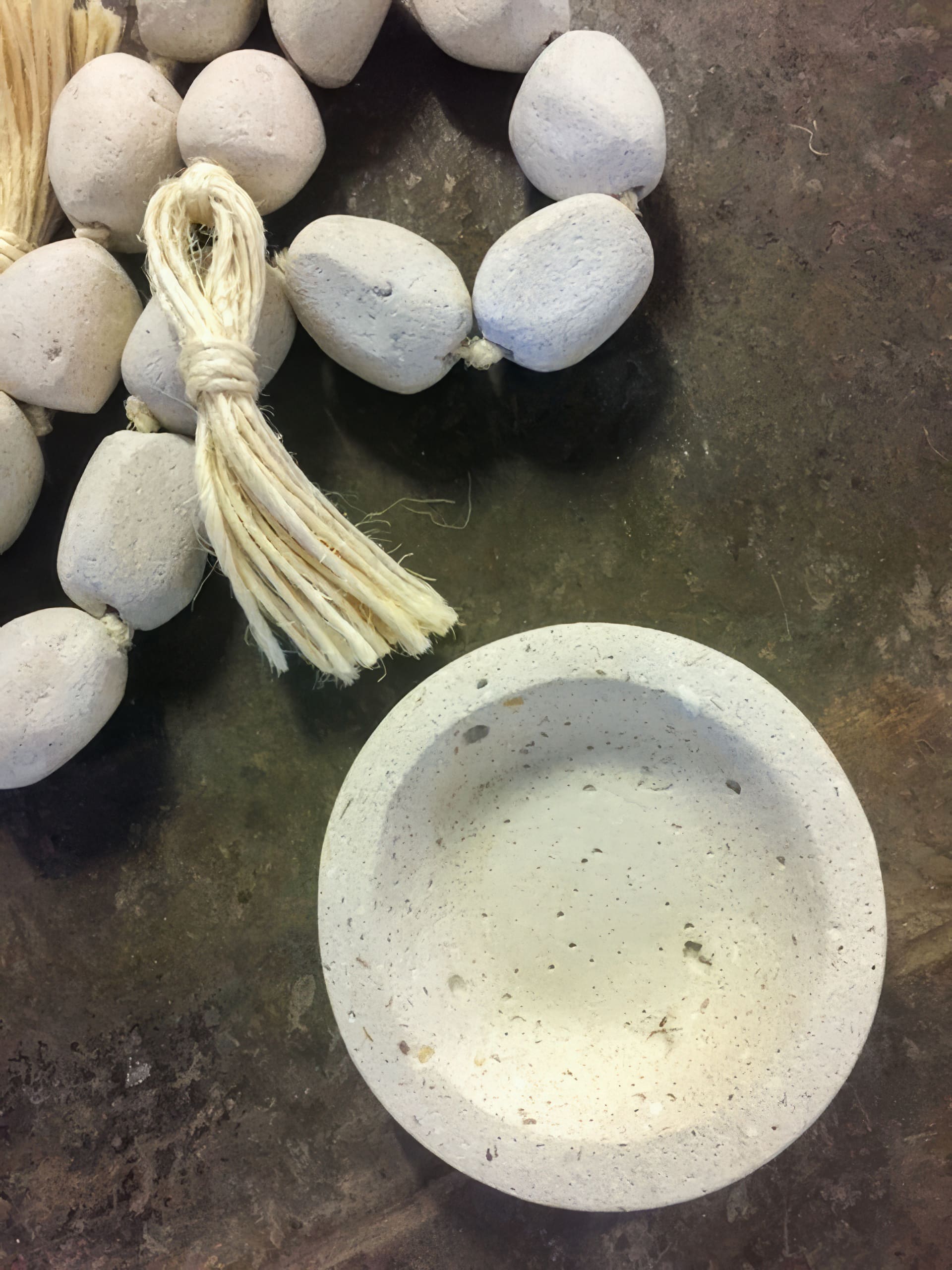 A round, shallow, textured white bowl sits on a mottled brown surface next to a string of white, oval beads with visible pores. A bundle of cream-coloured fibres is tied near the beads, giving a natural, handmade appearance.