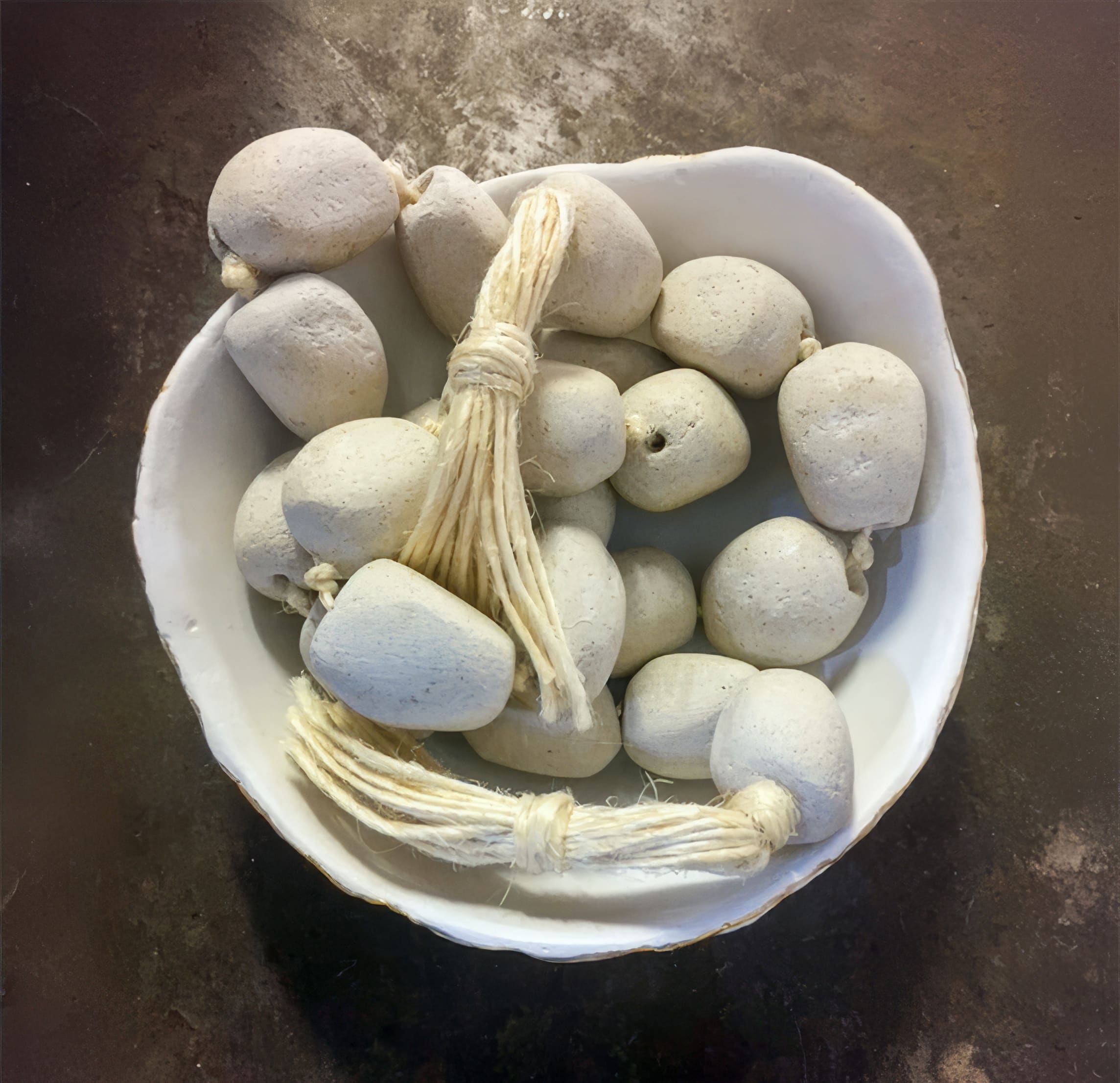 A white ceramic bowl filled with several white, irregularly shaped clay beads strung together with coarse natural twine, placed on a dark textured surface. Some beads are loose, whilst others are knotted in bunches.
