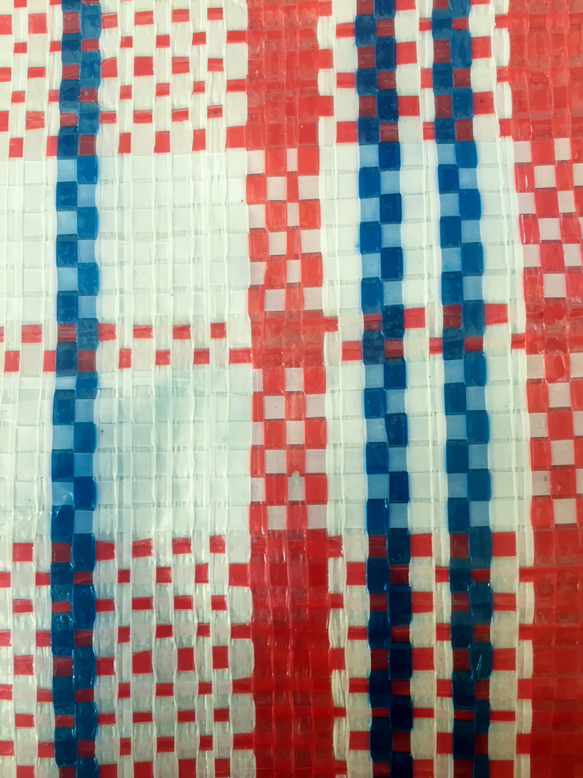 Close-up of a woven plastic material featuring a chequered pattern with red, white, and blue strips. The pattern alternates vertical and horizontal lines, creating small squares. The texture is glossy and slightly reflective, showing the weave detail.