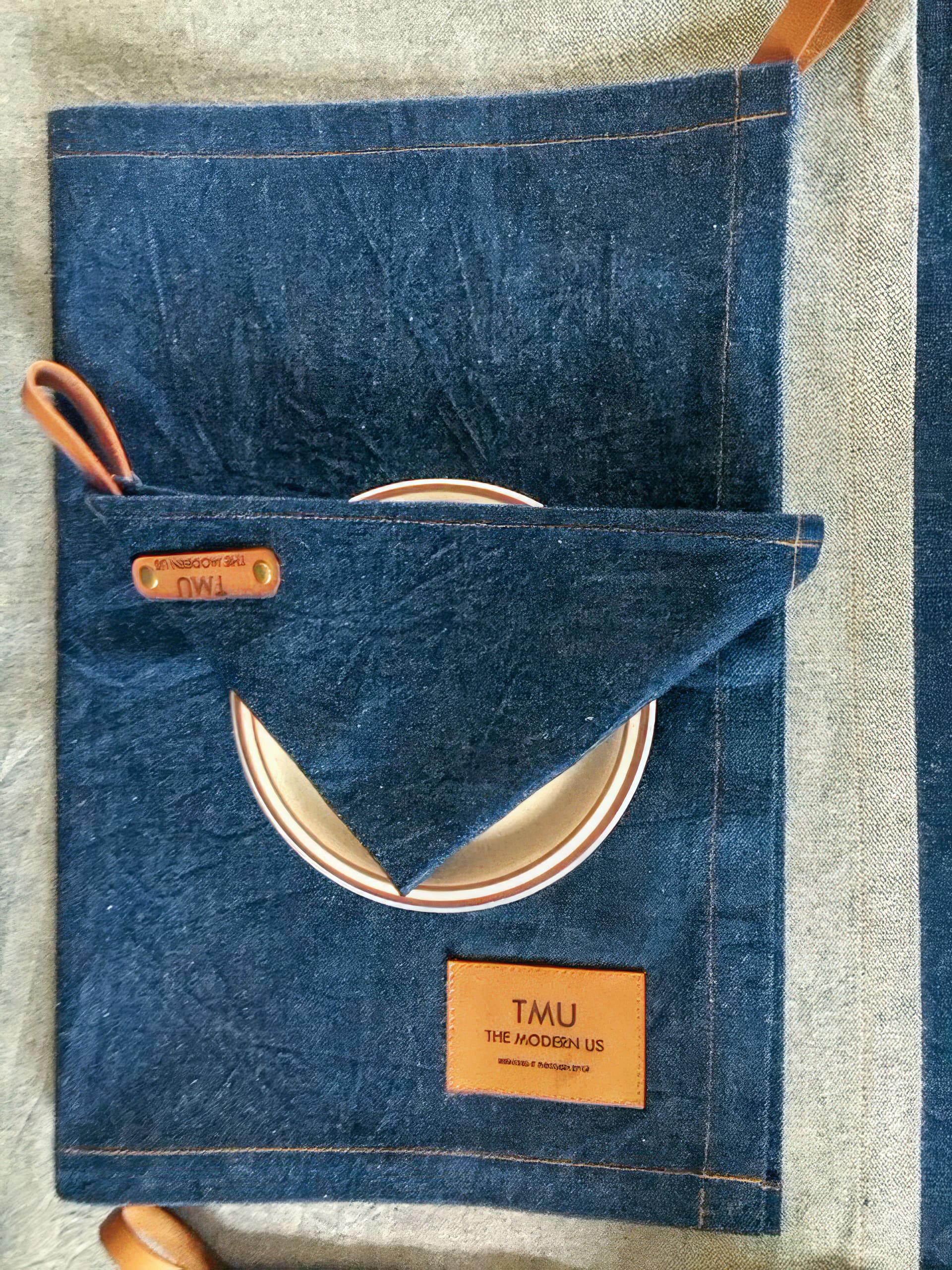 A folded dark blue denim placemat with brown leather accents and a label reading “TMU THE MODERN US.” A white plate with a brown rim is partially tucked into a triangular pocket on the placemat. The surface below is a lighter fabric.