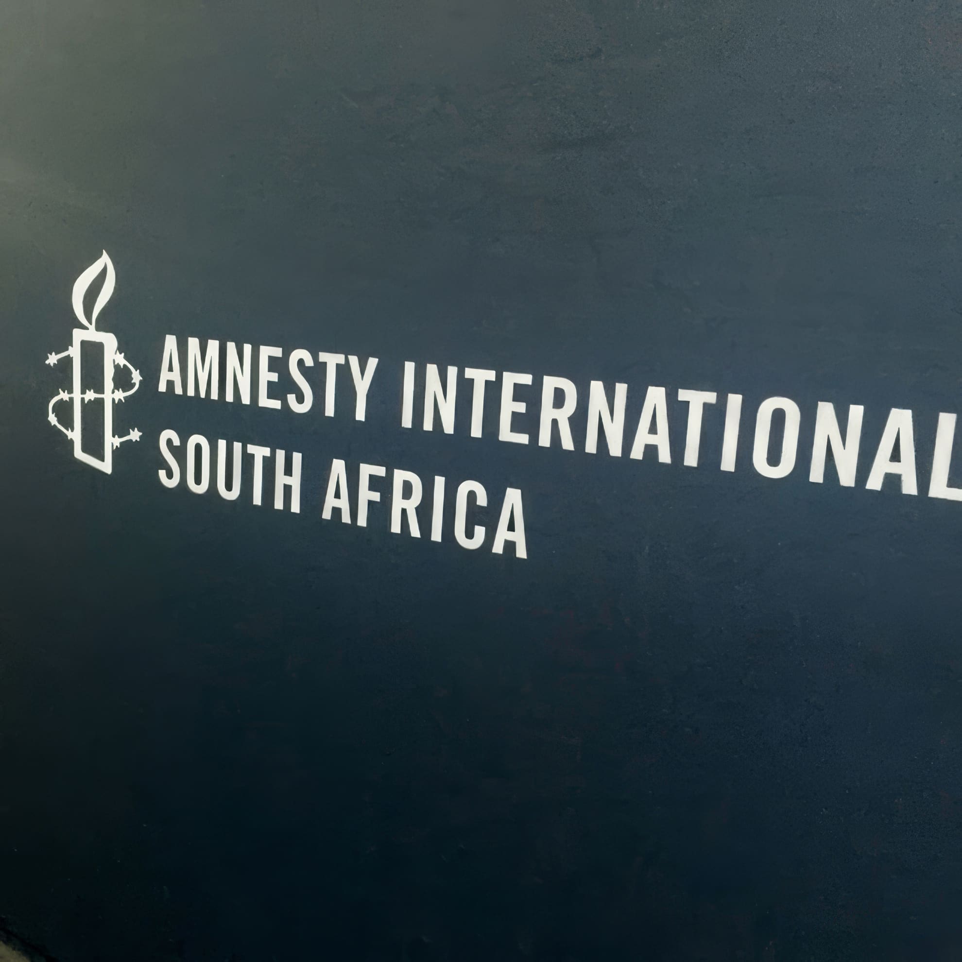 A dark wall displays the white logo of Amnesty International—a candle wrapped in barbed wire—next to bold text reading 
