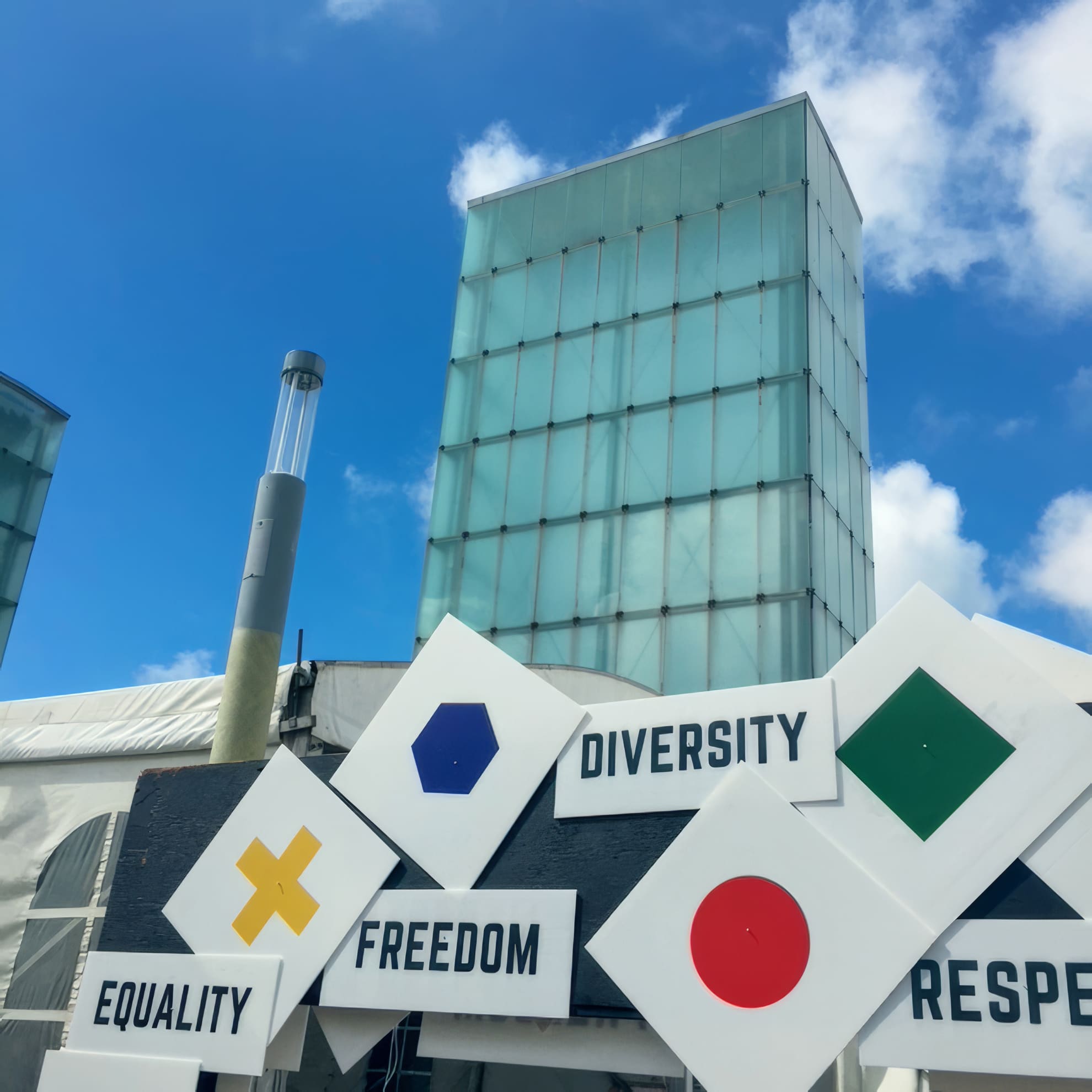 Modern glass buildings under a blue sky form the background. In the foreground, white geometric signs display words: 