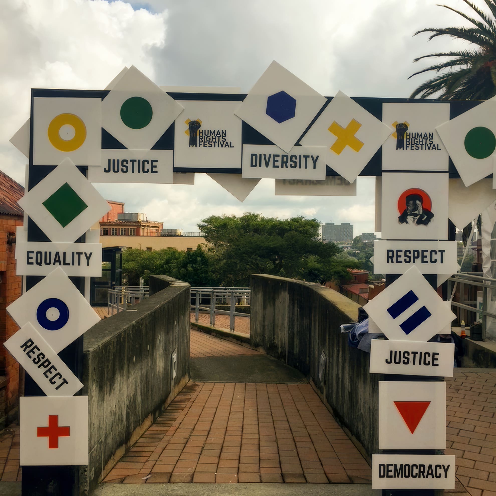 A festival archway displays bold geometric symbols and signs with words like “Justice,” “Diversity,” “Equality,” “Respect,” and “Democracy.” The sky is cloudy, trees and buildings are visible beyond the walkway, and the event logo reads “Human Rights Festival”.