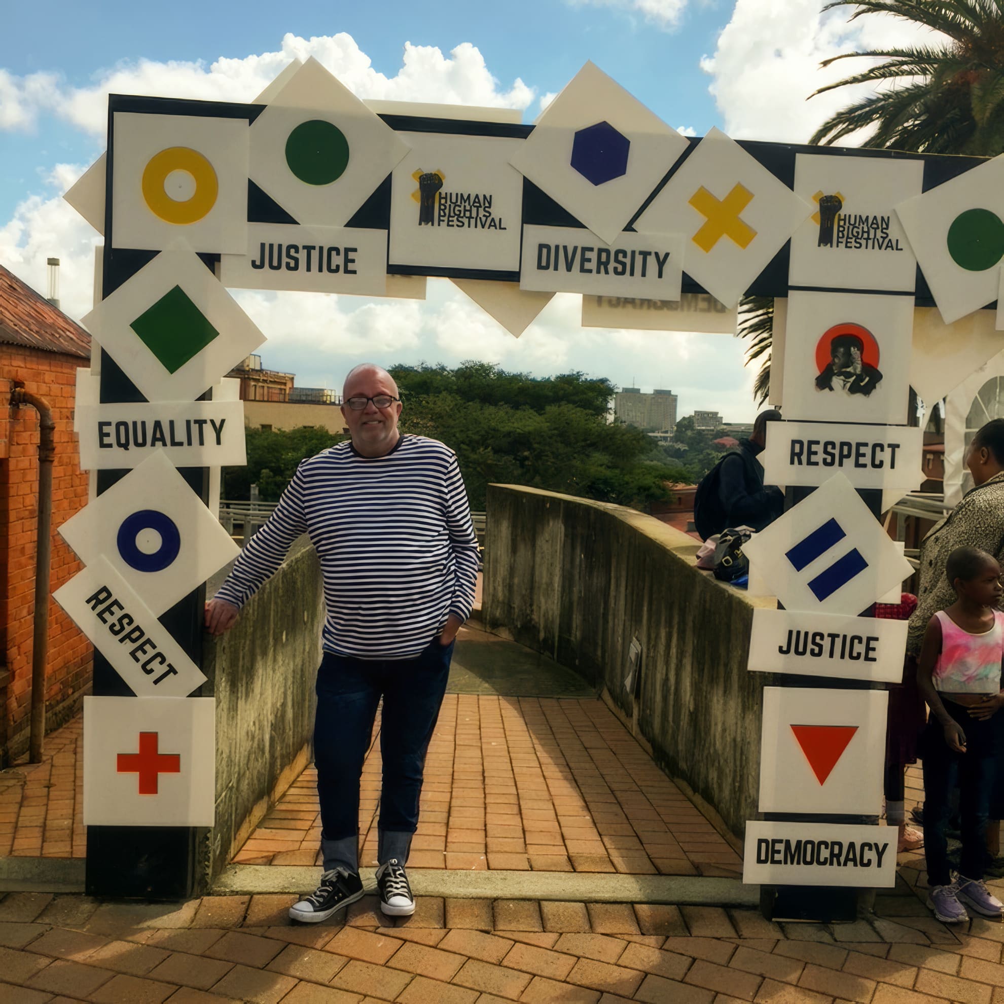 A man in glasses and a striped shirt stands under an outdoor archway decorated with geometric shapes and signs reading “Equality”, “Respect”, “Justice”, “Diversity”, and “Democracy”. Two children stand to the right. It’s a sunny day with scattered clouds.